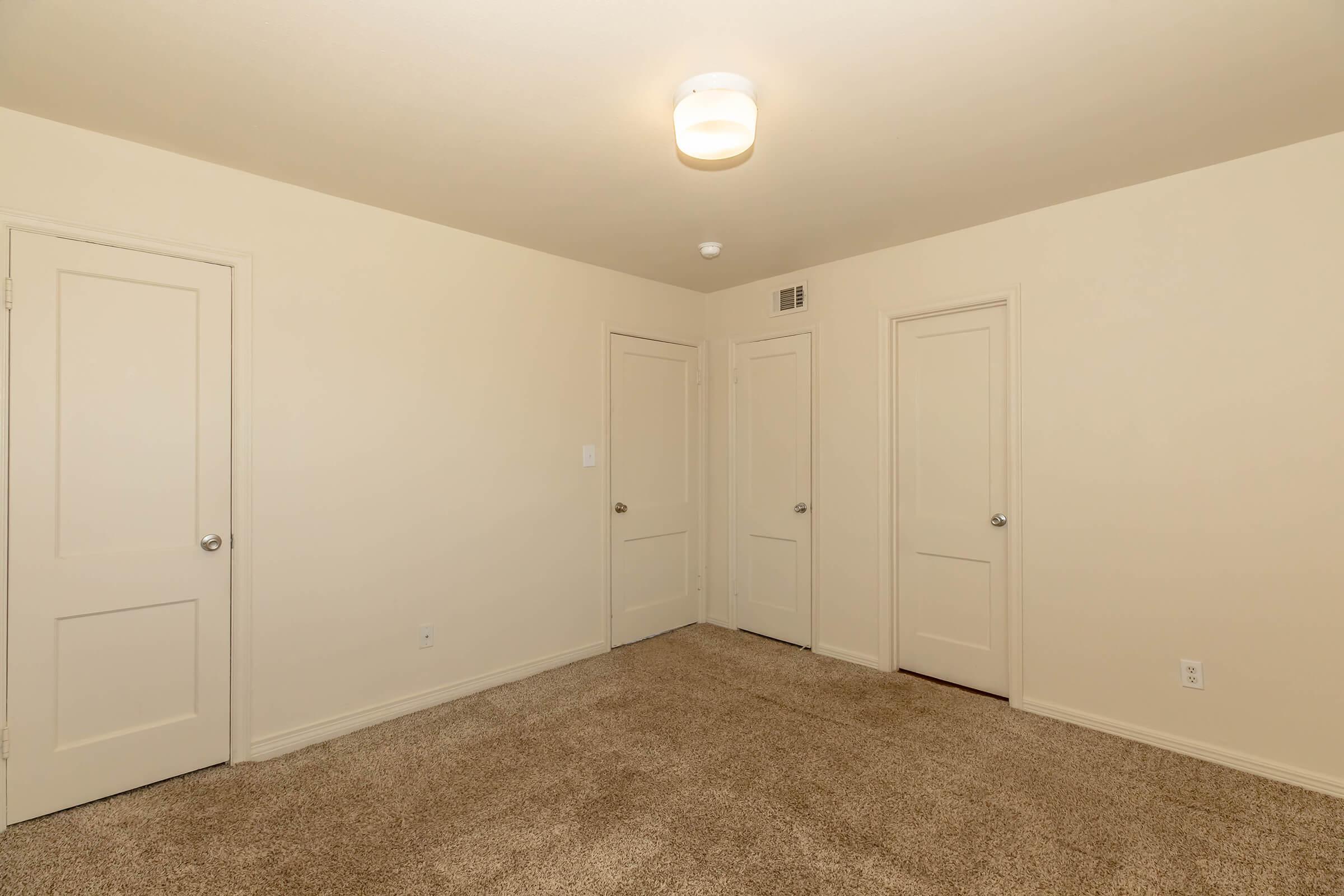 Empty room featuring beige walls and a warm light fixture on the ceiling. The carpeted floor is a light brown color. Three doors are visible on the walls, leading to different areas, with simple metal doorknobs. The room has a spacious feel, with no furniture or decorations.