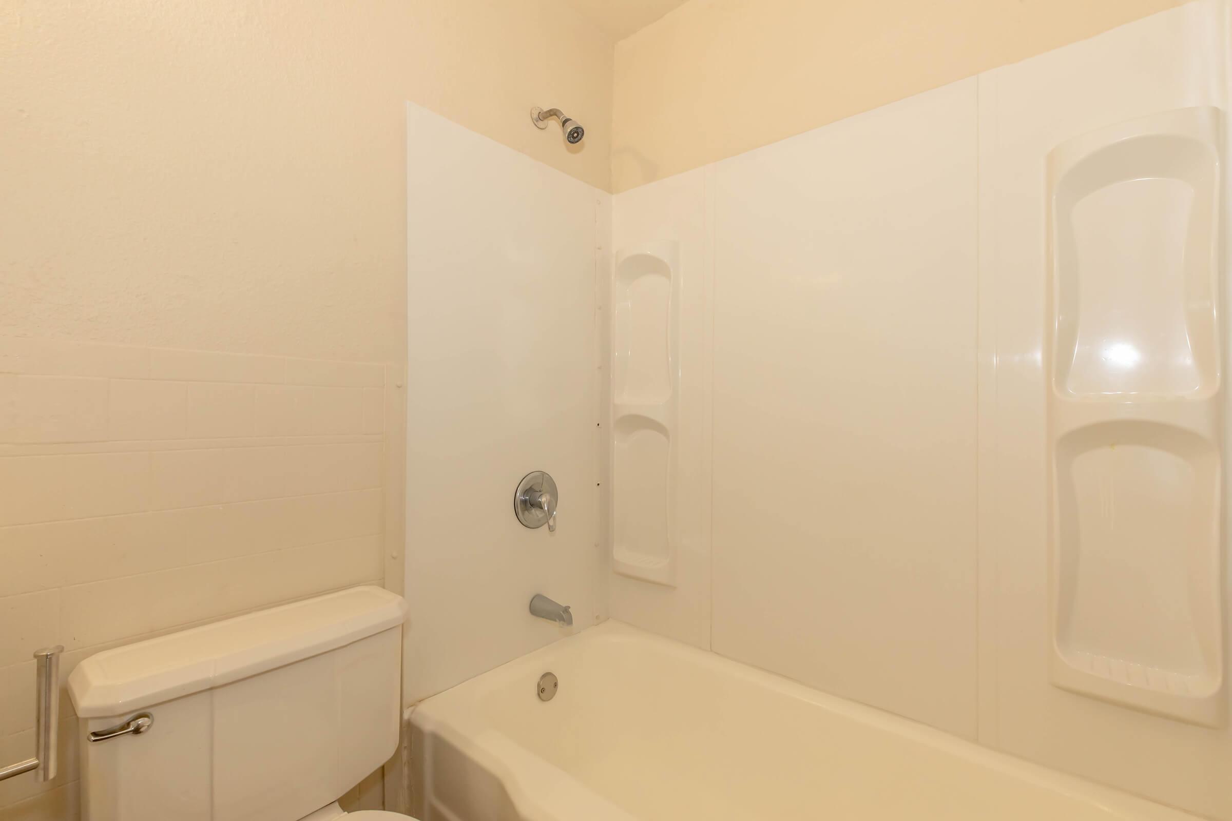 A clean, well-lit bathroom featuring a white bathtub with a showerhead overhead. The tub is surrounded by white wall panels, and there are two built-in shelves for toiletries. A toilet is visible beside the bathtub. The overall look is simple and modern.