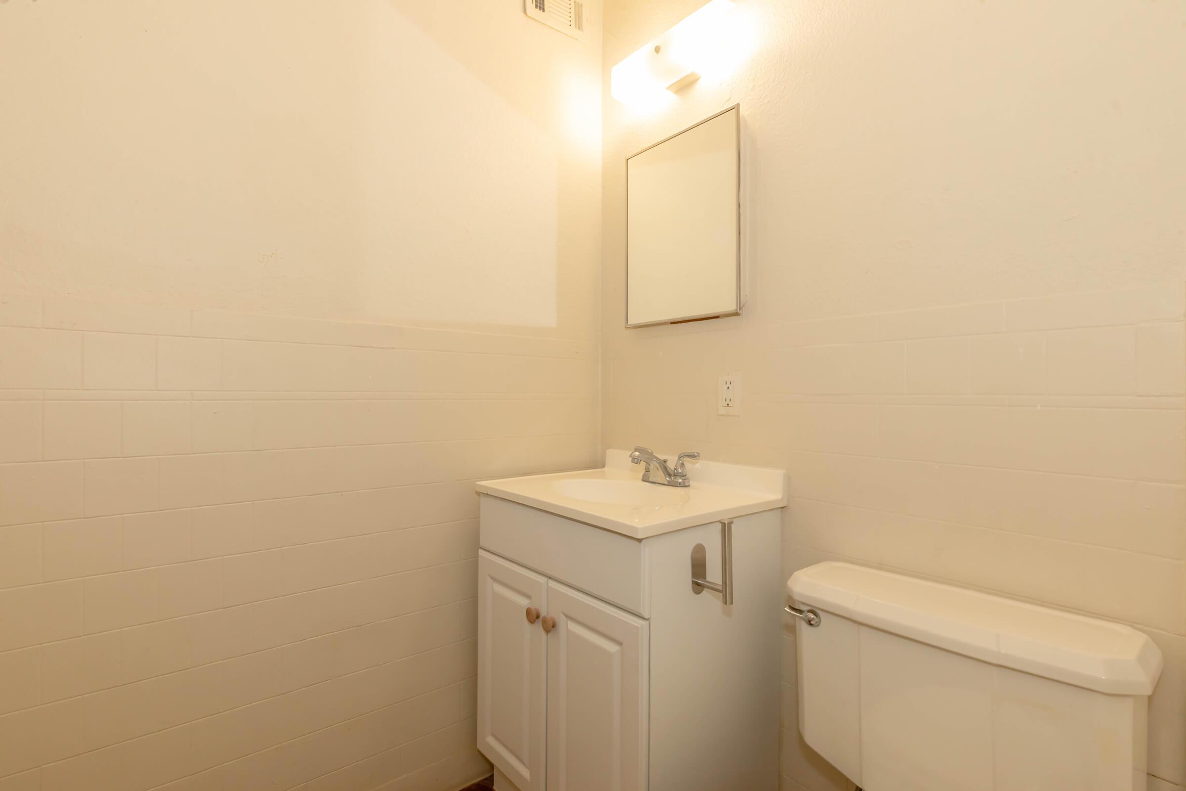 A clean, minimalist bathroom featuring a white vanity with a sink and a mirror above it. The wall behind is painted a light color, and there’s a toilet beside the vanity. Soft lighting is provided by a fixture above the mirror, creating a bright and airy atmosphere.