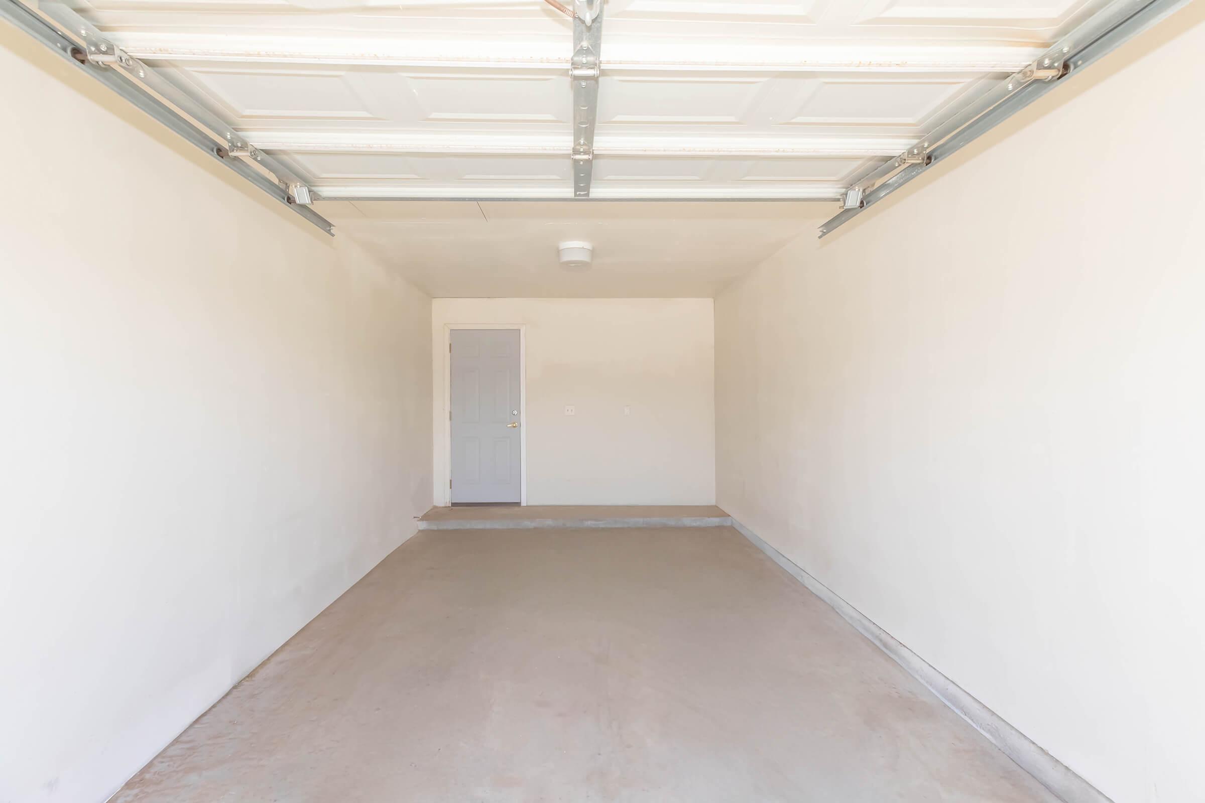 An empty garage with white walls, a concrete floor, and a closed garage door. A single gray door is visible on the left wall. The space is well-lit and free of any furniture or items, creating a clean and open appearance.