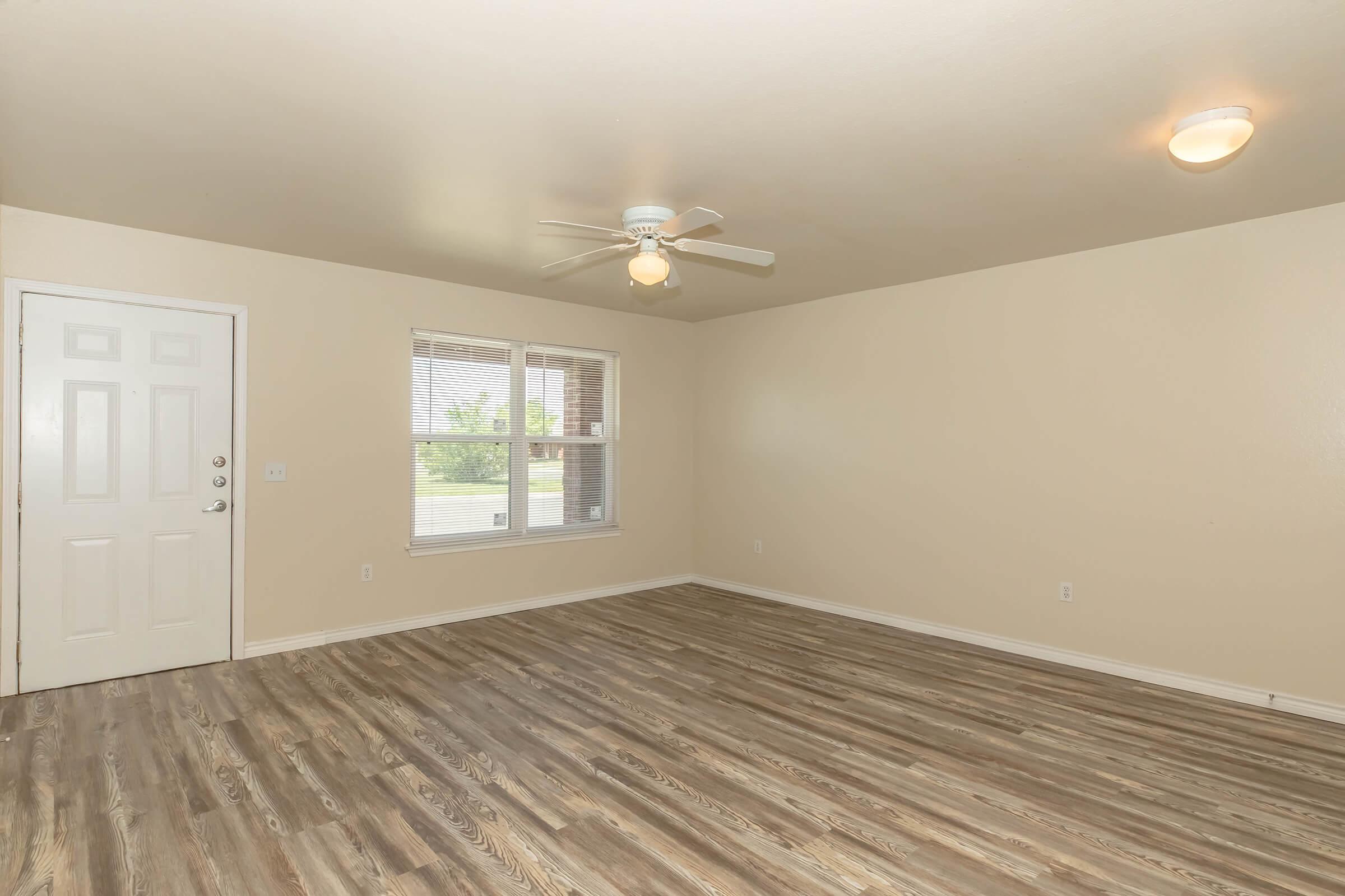 Empty room with light-colored walls and a ceiling fan. A single door is visible on the left, along with a window featuring blinds. The flooring is a wood-like laminate, and the space is well-lit with a ceiling light. There are no furnishings or decorations, creating a clean, open appearance.