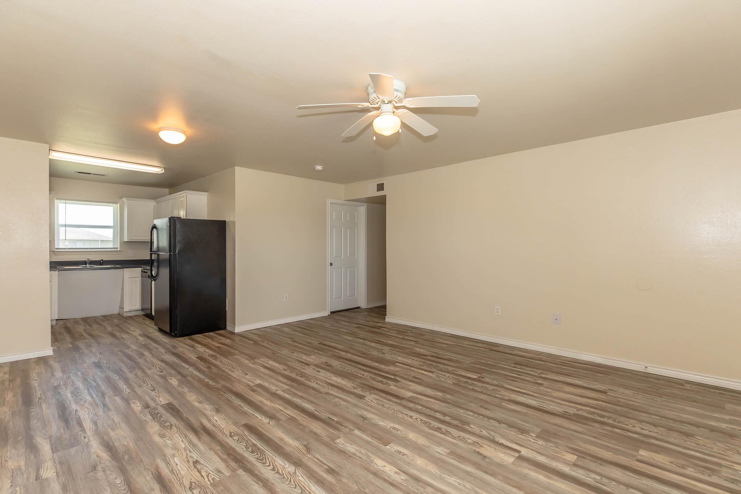 A spacious, empty room featuring light-colored walls and wooden flooring, with a ceiling fan and overhead lighting. In the background, a kitchen area is visible, equipped with white cabinets and a black refrigerator. The layout suggests an open concept design.