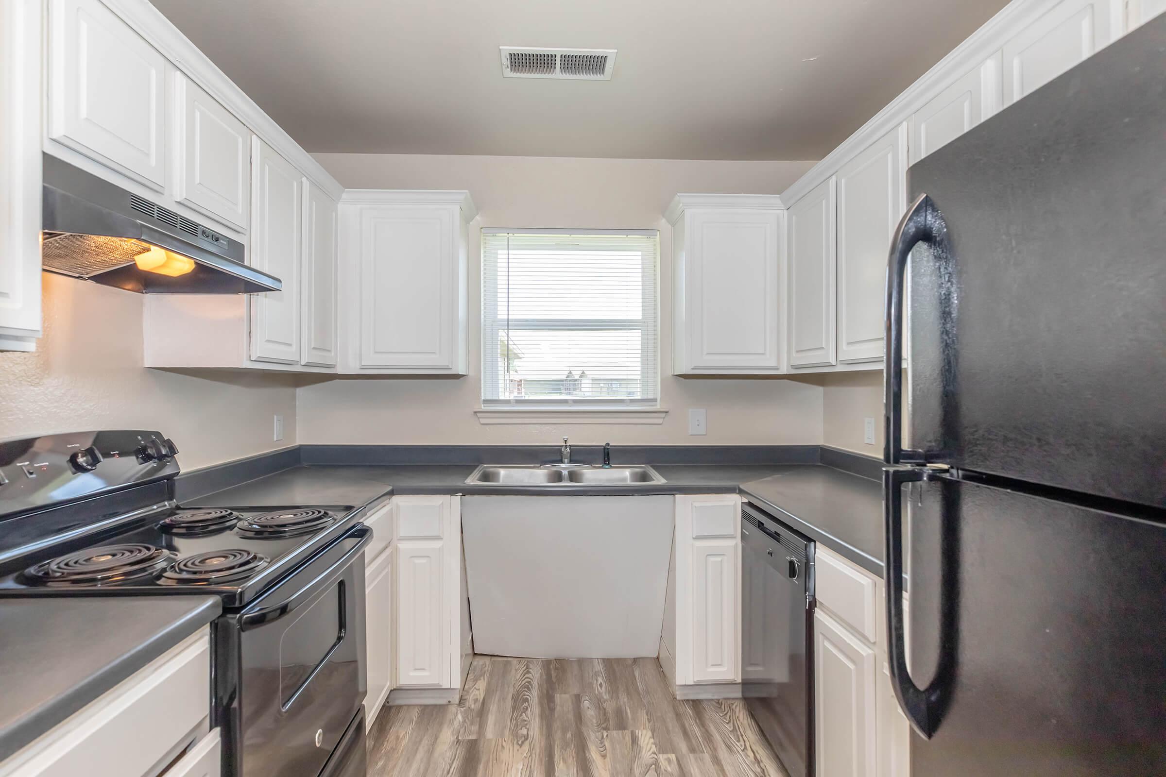 A modern kitchen featuring white cabinets, a black refrigerator, a black stove, and a double sink beneath a window. The countertops are dark, complementing the light-colored walls, and the flooring is a light wood finish. The kitchen is bright and functional, designed for easy use.