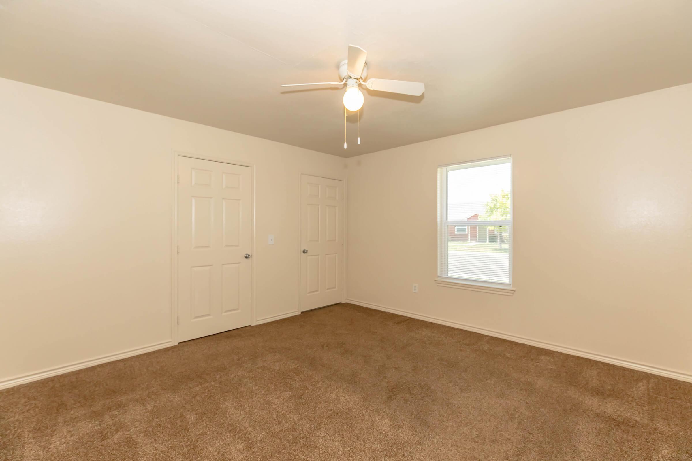 A spacious, empty room with light beige walls and carpet. Two white doors are visible, one on the left and one on the right, leading to closets. A ceiling fan is mounted in the center, and a window allows natural light to enter, illuminating the space. The room feels bright and inviting.