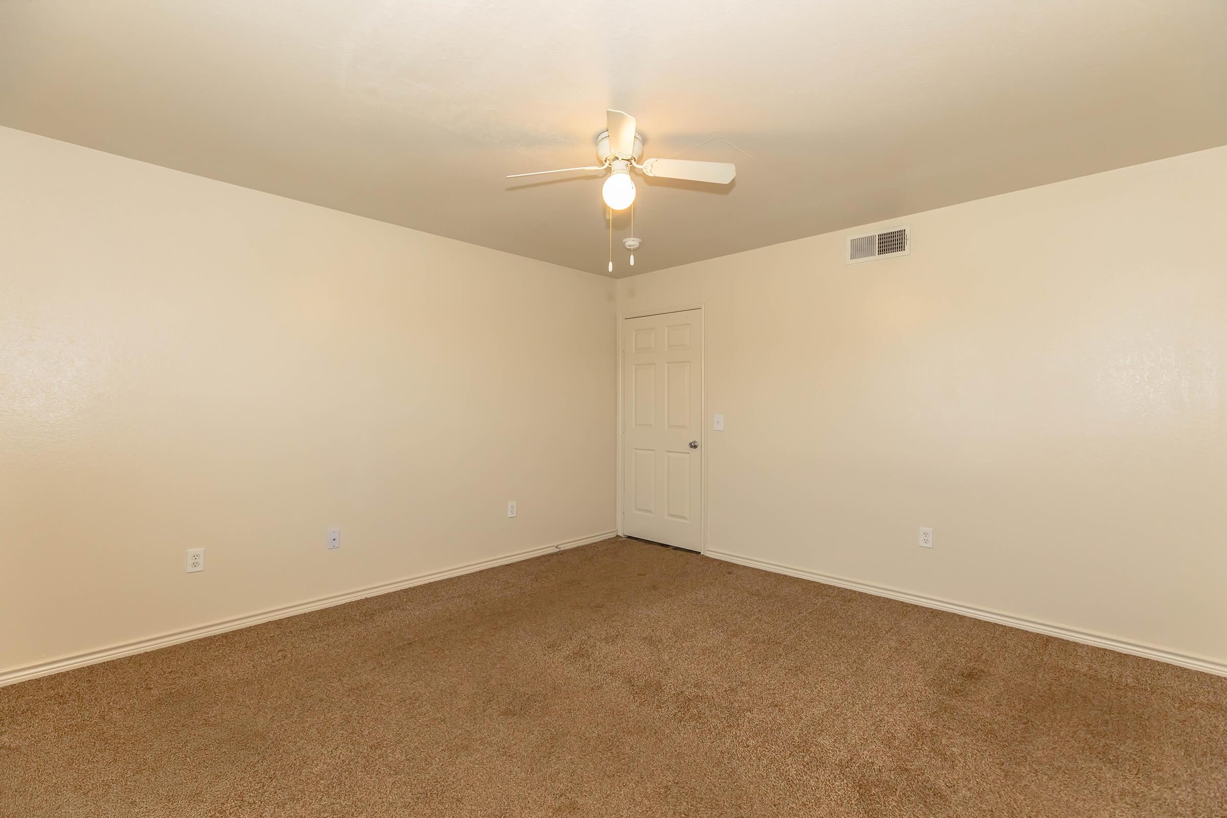 Empty room with beige walls and a light brown carpet. The ceiling features a white ceiling fan, and there is a closed door on the right. The space is well-lit, with no furniture or decorations, creating a clean and open atmosphere.