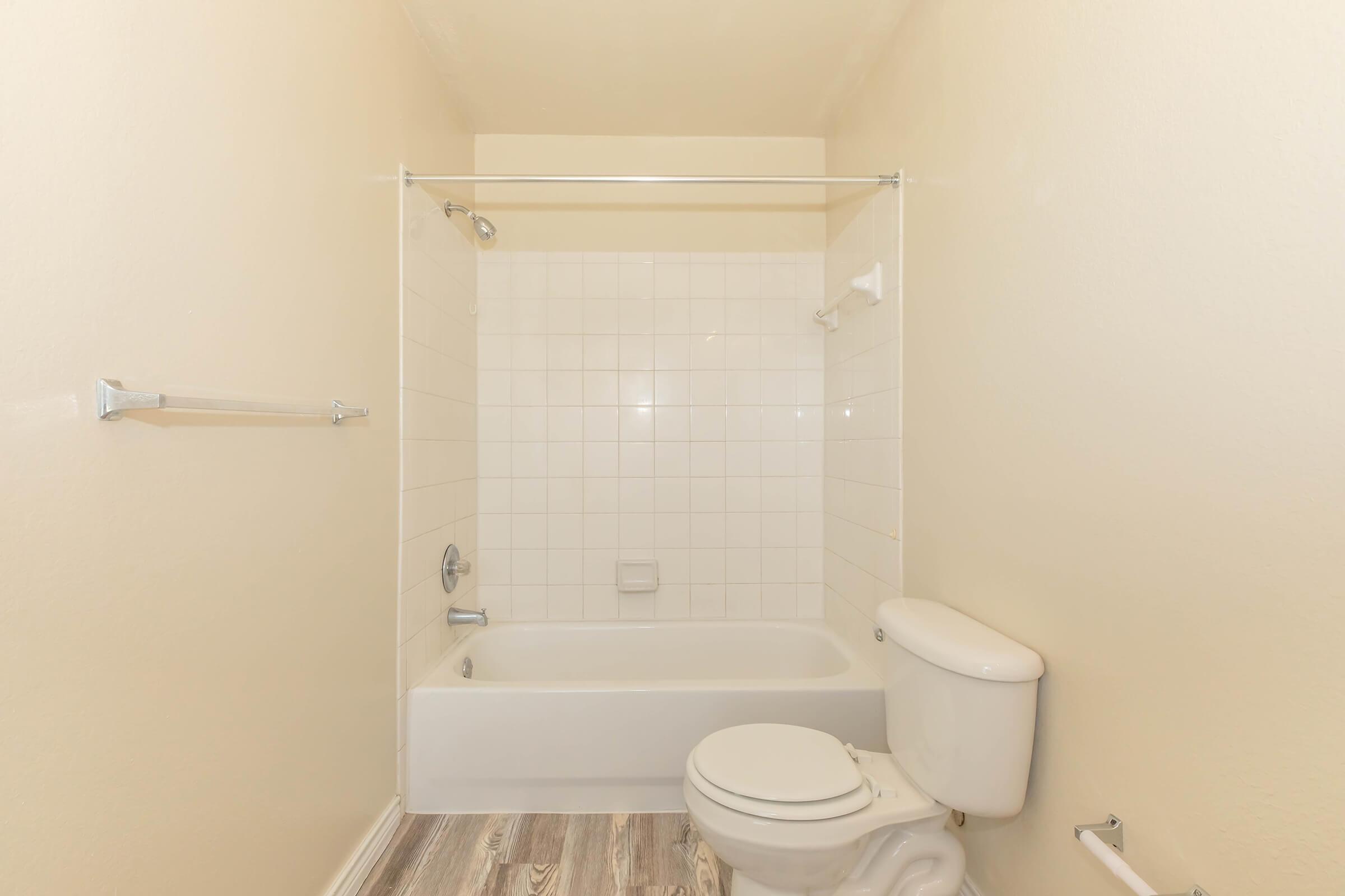 Empty bathroom featuring a shower-tub combo with white tiles, a toilet, and a small window. The walls are painted a light beige color, and the floor has light wood-like tiles. There are grab bars and a towel rack visible on the walls.