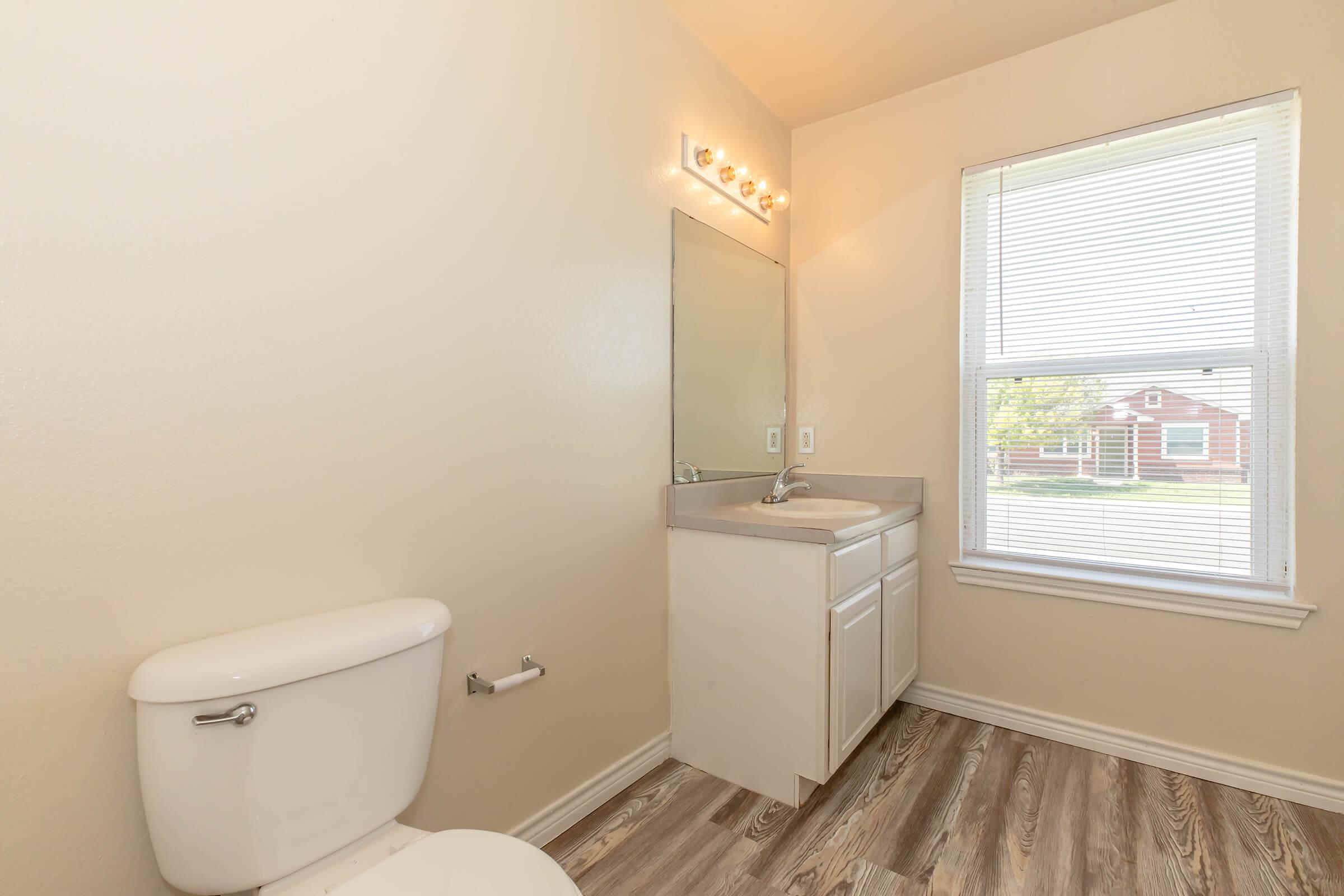 A bright and clean bathroom featuring a white toilet, a mirror above a sink with a gray countertop, and natural light coming through a window with blinds. The floor is covered in light wood-like flooring, and the walls are painted in a neutral color.
