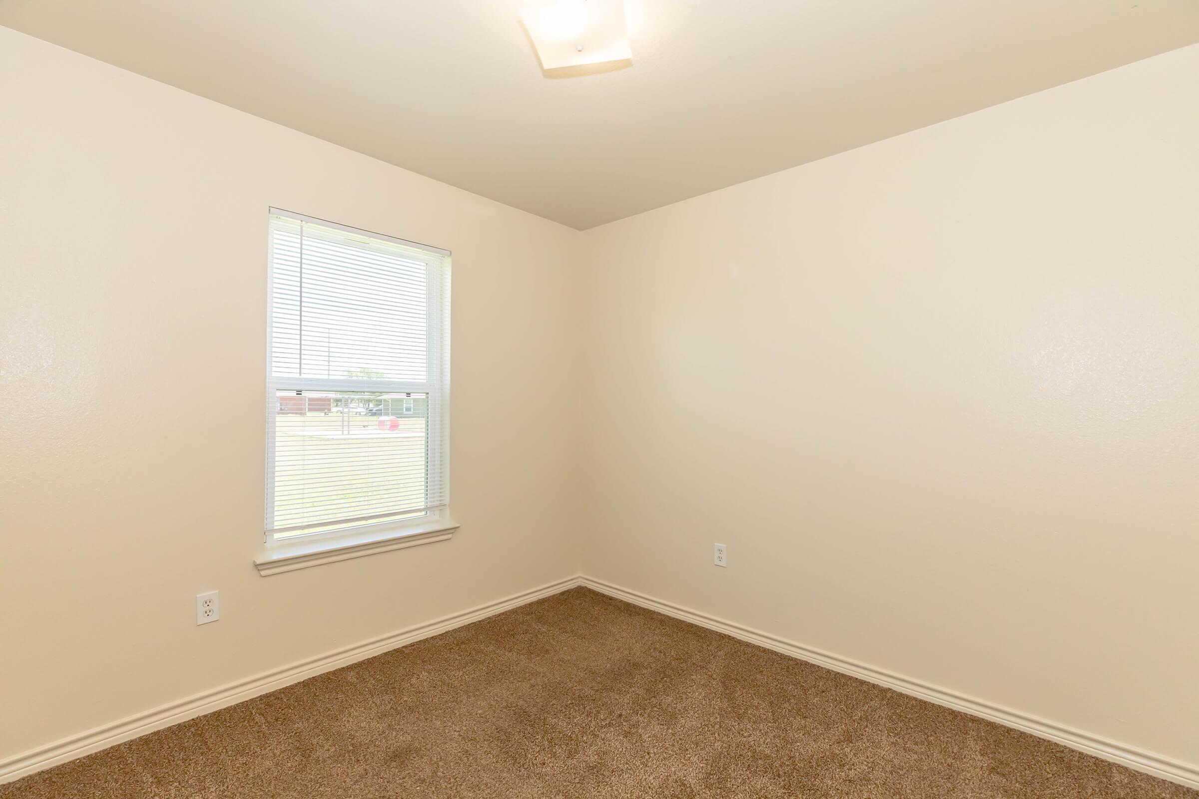 A small, light-colored room with beige carpet and plain walls. There is a single window with white blinds allowing natural light to enter. The ceiling has a light fixture, and the room appears empty, giving it a clean and spacious feel.