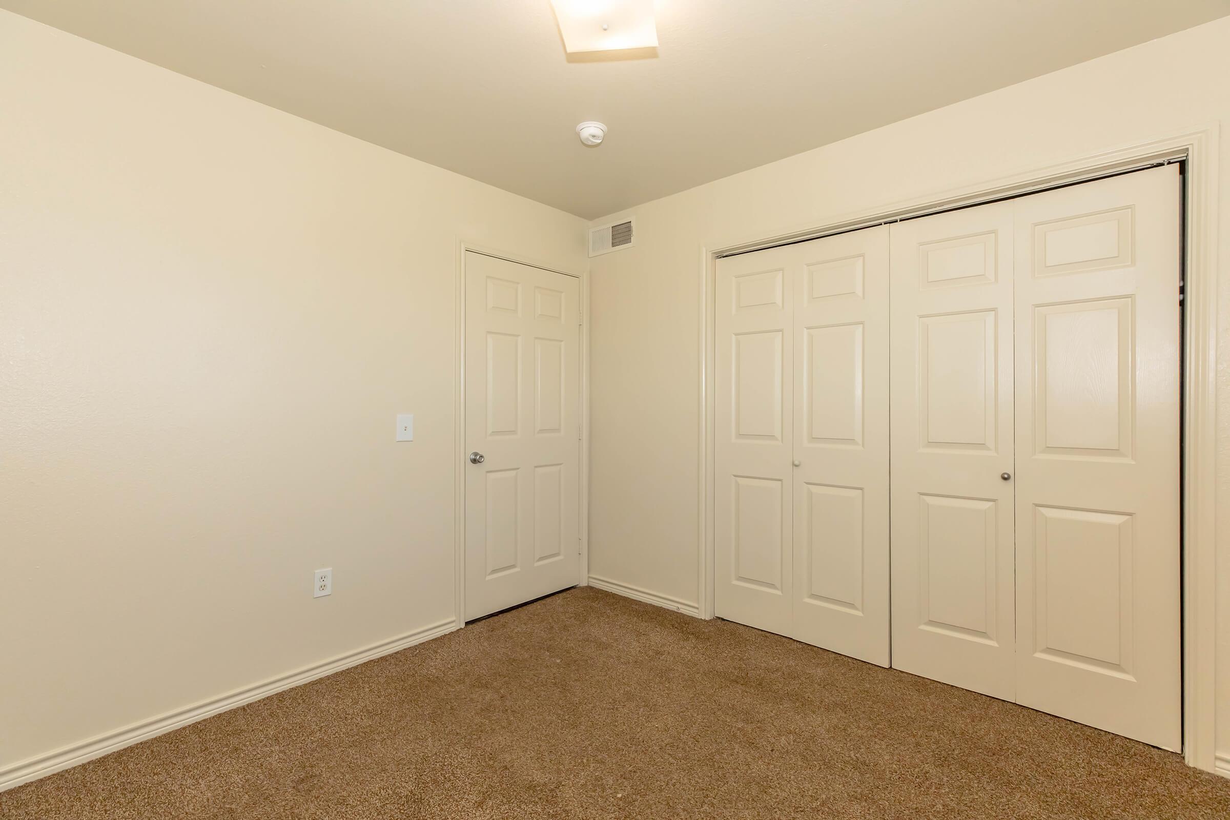 A spacious, empty room with light beige walls and carpeted flooring. There are two closed doors: one leading to the hallway and the other with double panels for a closet. A ceiling light fixture provides illumination, enhancing the room's bright and simple appearance.