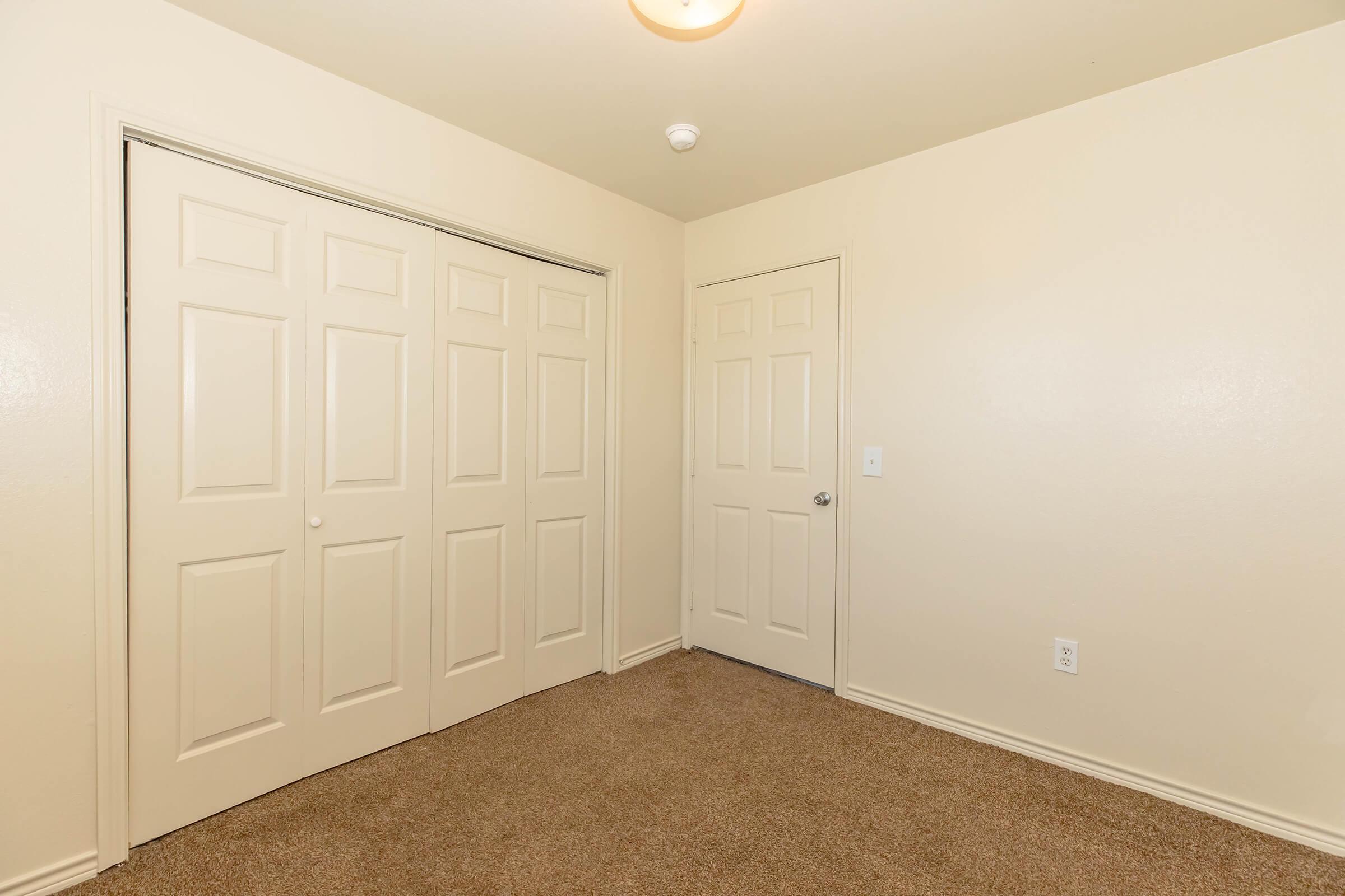 A simple room featuring beige walls and carpeted flooring. Two closed white panel doors are visible: one leads to a closet, and the other is a standard door. There is a light fixture on the ceiling, adding brightness to the space.