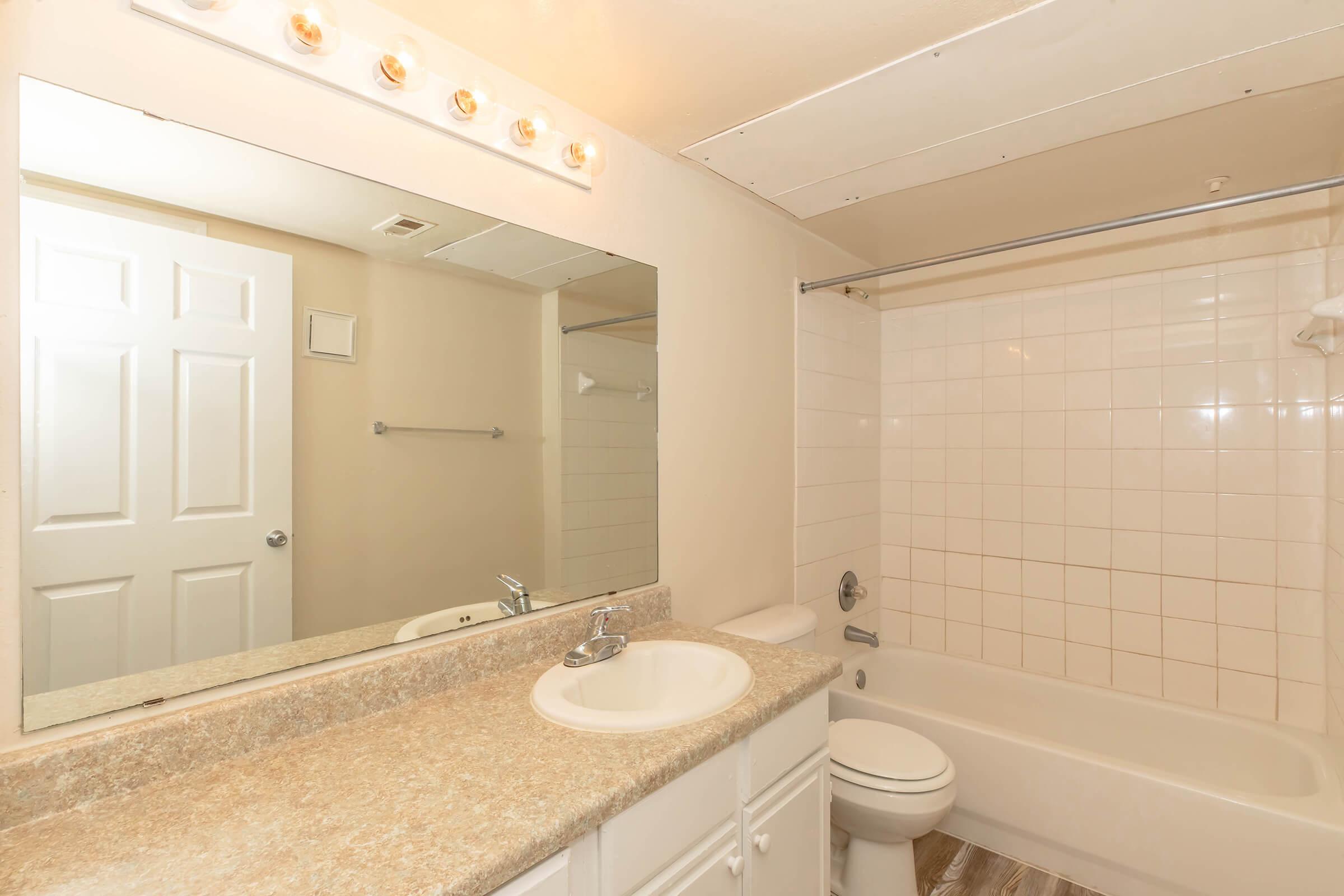 A spacious bathroom featuring a large mirror above a sink with a countertop, a bathtub with a shower curtain, and a white toilet. The walls are tiled, and there are overhead lights. A door is partially visible, indicating access to another room.