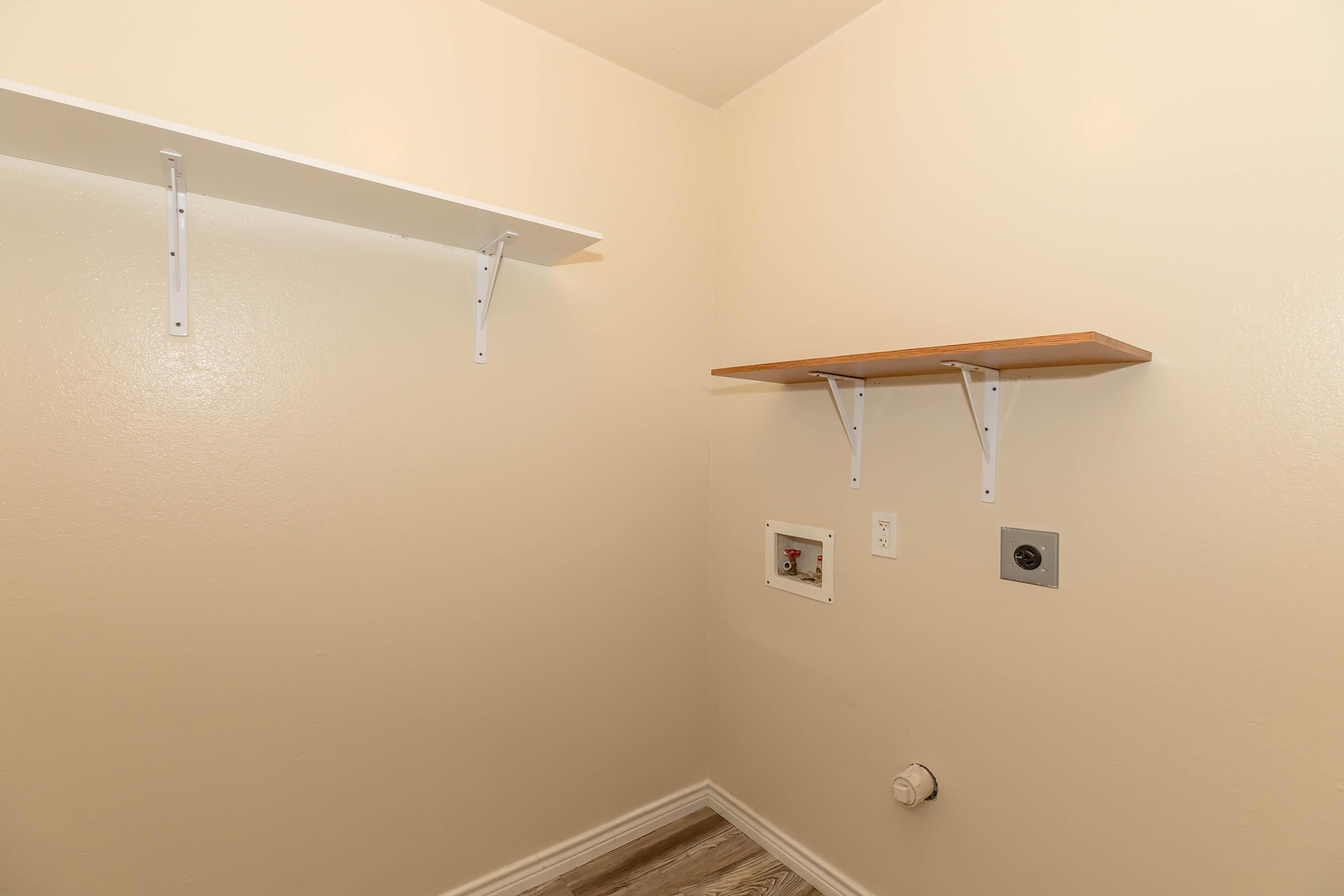 Empty corner of a room featuring two shelves—one white and one wooden—attached to beige walls. There are electrical outlets and a small utility panel visible. The flooring appears to be light-colored wood or laminate. The space is well-lit and minimalistic.