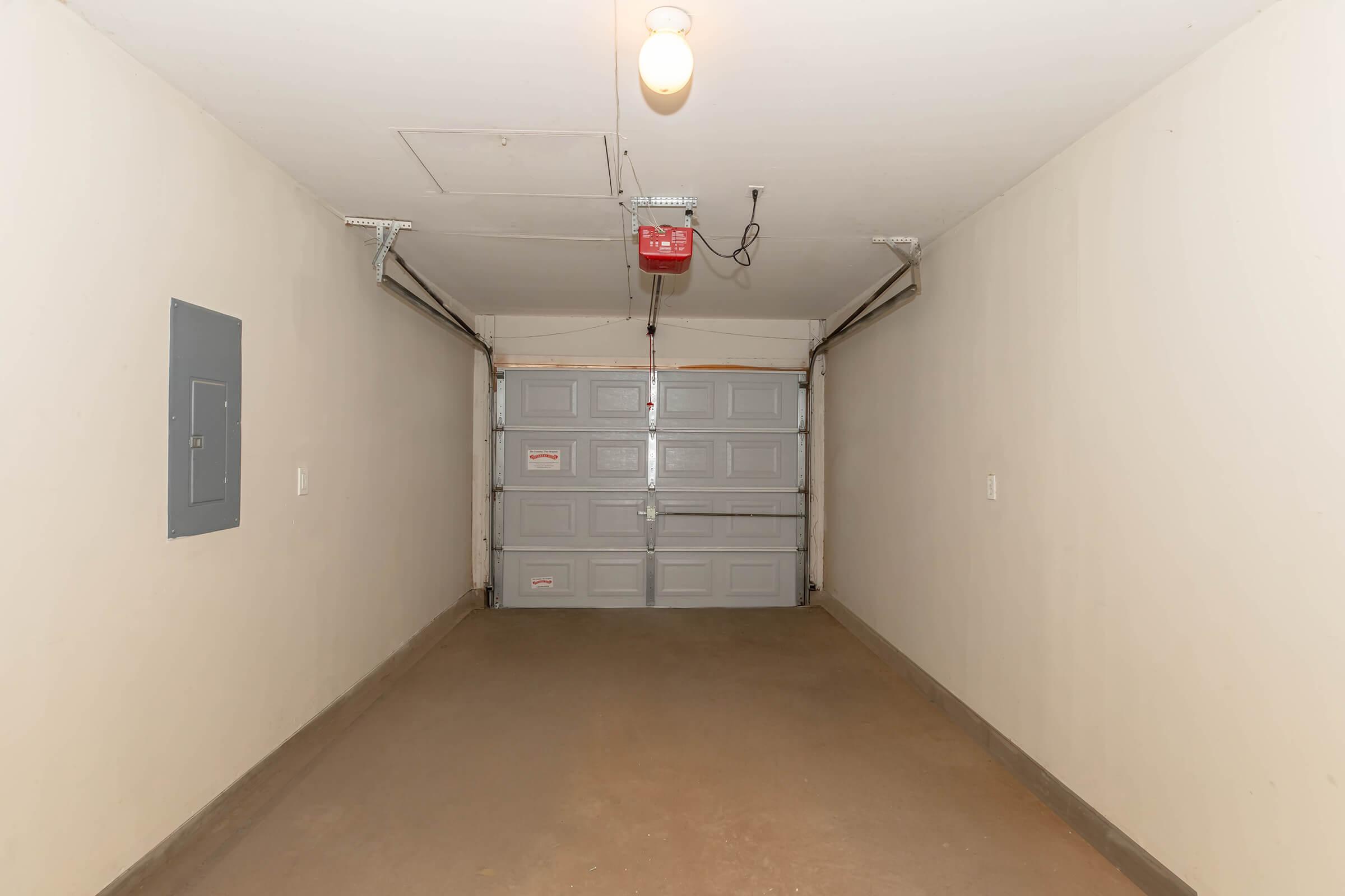 Interior view of a spacious garage, featuring a concrete floor, plain beige walls, a ceiling light, and a garage door with a red opener. The garage is empty, showcasing ample storage space. There's an electrical panel on the left wall.