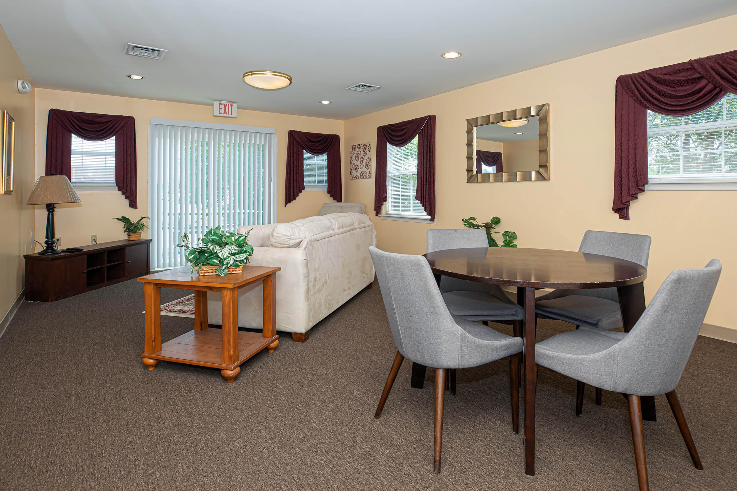 A cozy living space featuring a light-colored sofa, a wooden coffee table, and a round dining table with gray chairs. The walls are painted in a warm tone, and there are decorative curtains and plants enhancing the inviting atmosphere. Natural light comes through a sliding door.