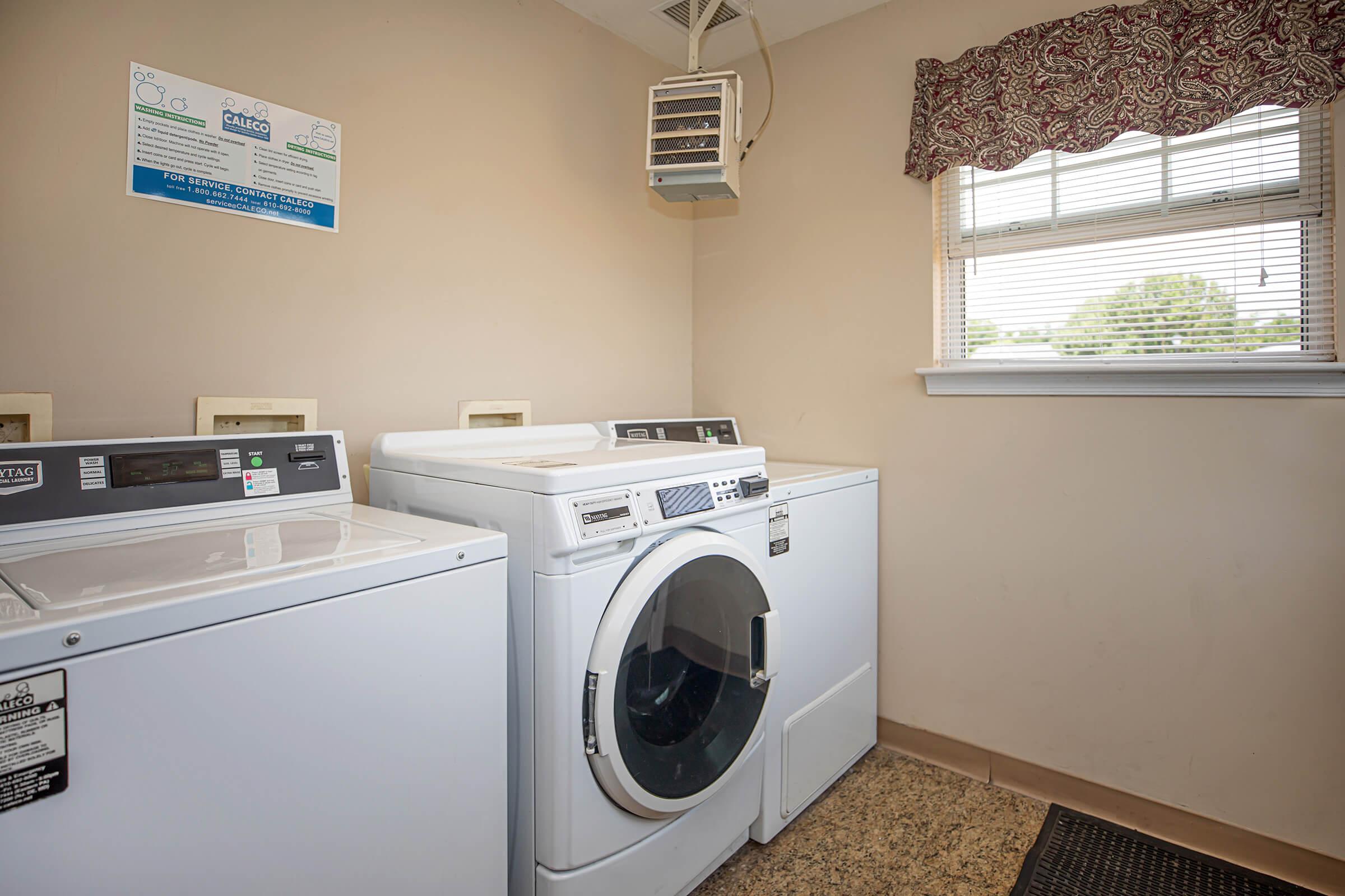 A laundry room featuring two washing machines and a dryer, with a window adorned with a patterned valance. There is also a wall-mounted drying unit and a notice on the wall providing information about the laundry facilities. The floor is finished in a light-colored tile.