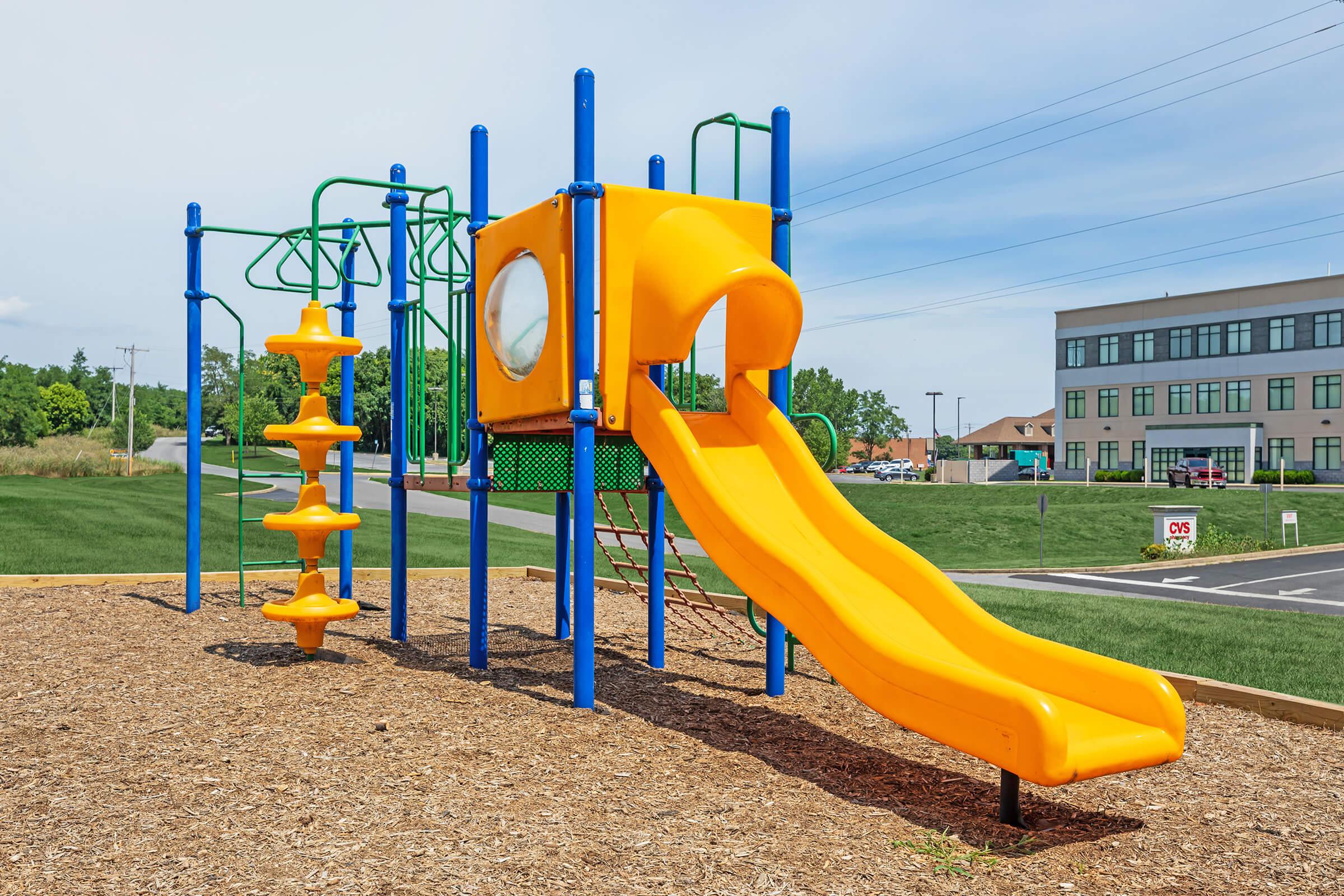 Colorful playground structure featuring a vibrant orange slide, a climbing frame with various climbing elements, and a transparent window. The structure is set on a bed of wood chips, surrounded by grassy areas and a building in the background. Clear blue sky overhead.