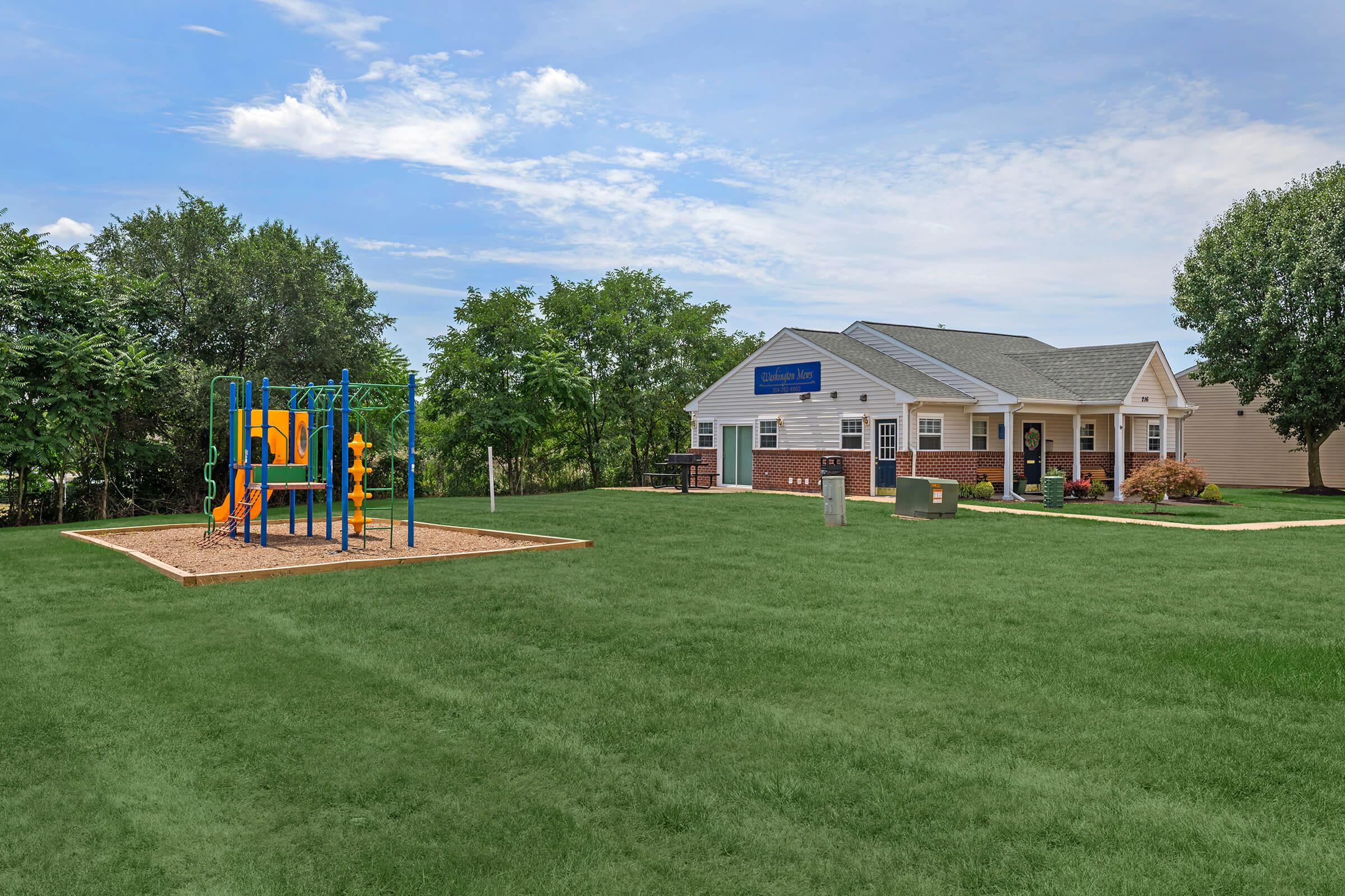 A colorful playground with a slide and climbing equipment sits on a grassy area beside a modern building. The building has a sign and features a sloped roof, surrounded by trees and open space under a partly cloudy sky.