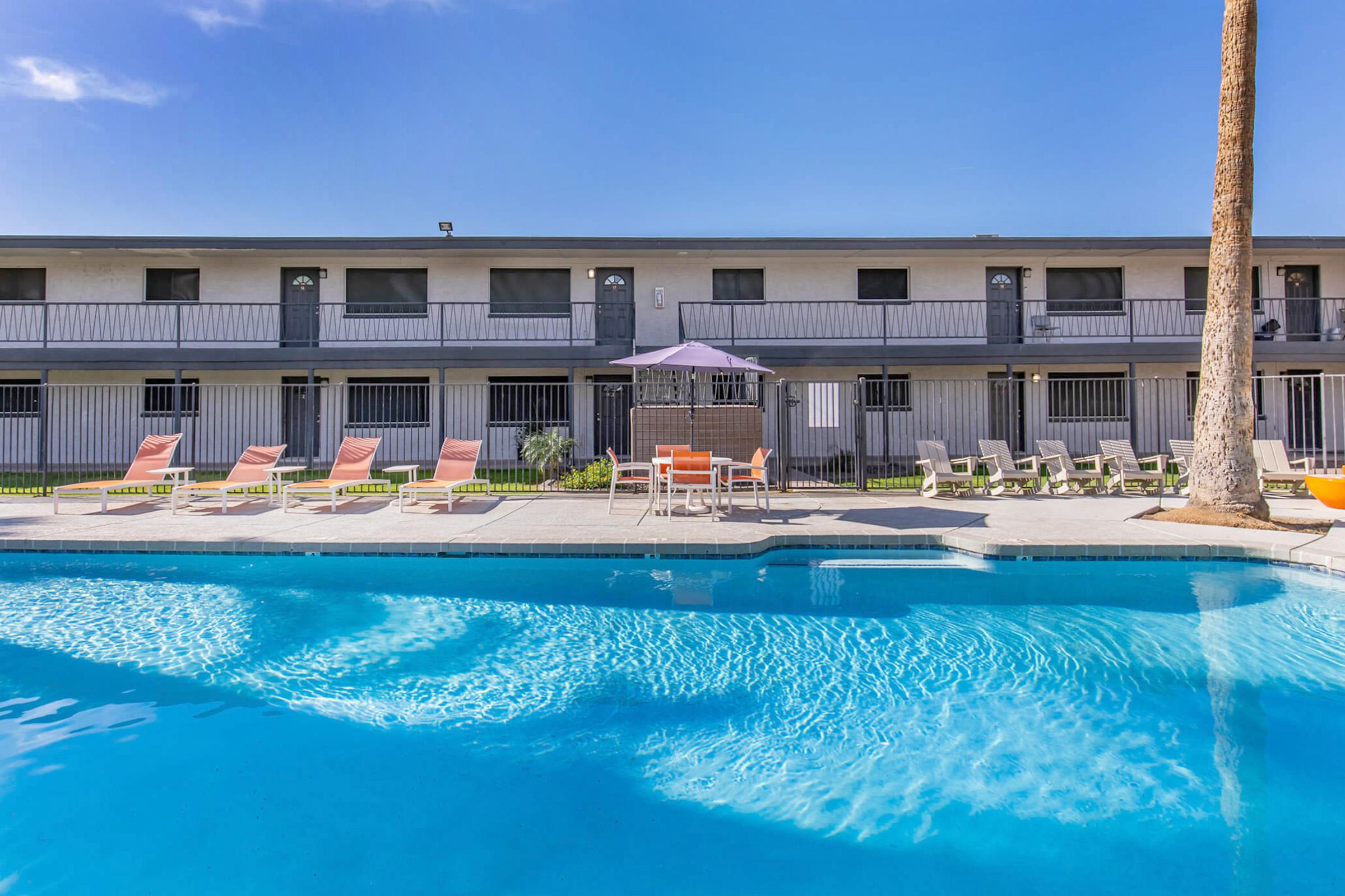 A clear blue swimming pool is in the foreground, with lounge chairs and a shaded table nearby. In the background, a two-story building with multiple rooms features balconies. The scene is set under a bright blue sky, suggesting a warm, sunny day.