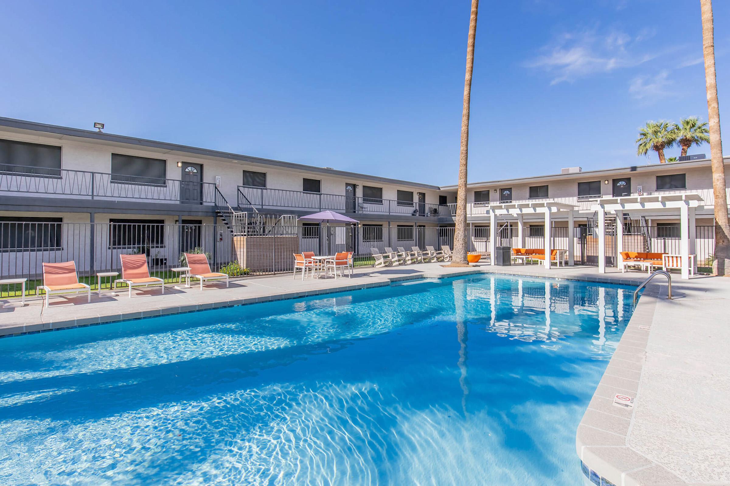 A view of a swimming pool surrounded by lounge chairs at a residential complex. The pool area features a shaded sitting area with umbrellas and is bordered by two palm trees. In the background, there's a two-story building with balconies and multiple windows under a clear blue sky.