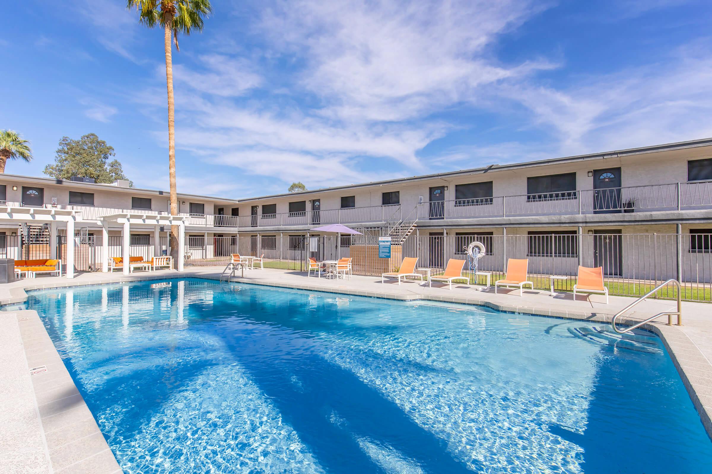 A clear blue swimming pool surrounded by orange lounge chairs, with a tall palm tree nearby. In the background, a two-story building with multiple balconies and windows under a bright blue sky with scattered clouds. The scene evokes a relaxed, sunny atmosphere perfect for leisure.