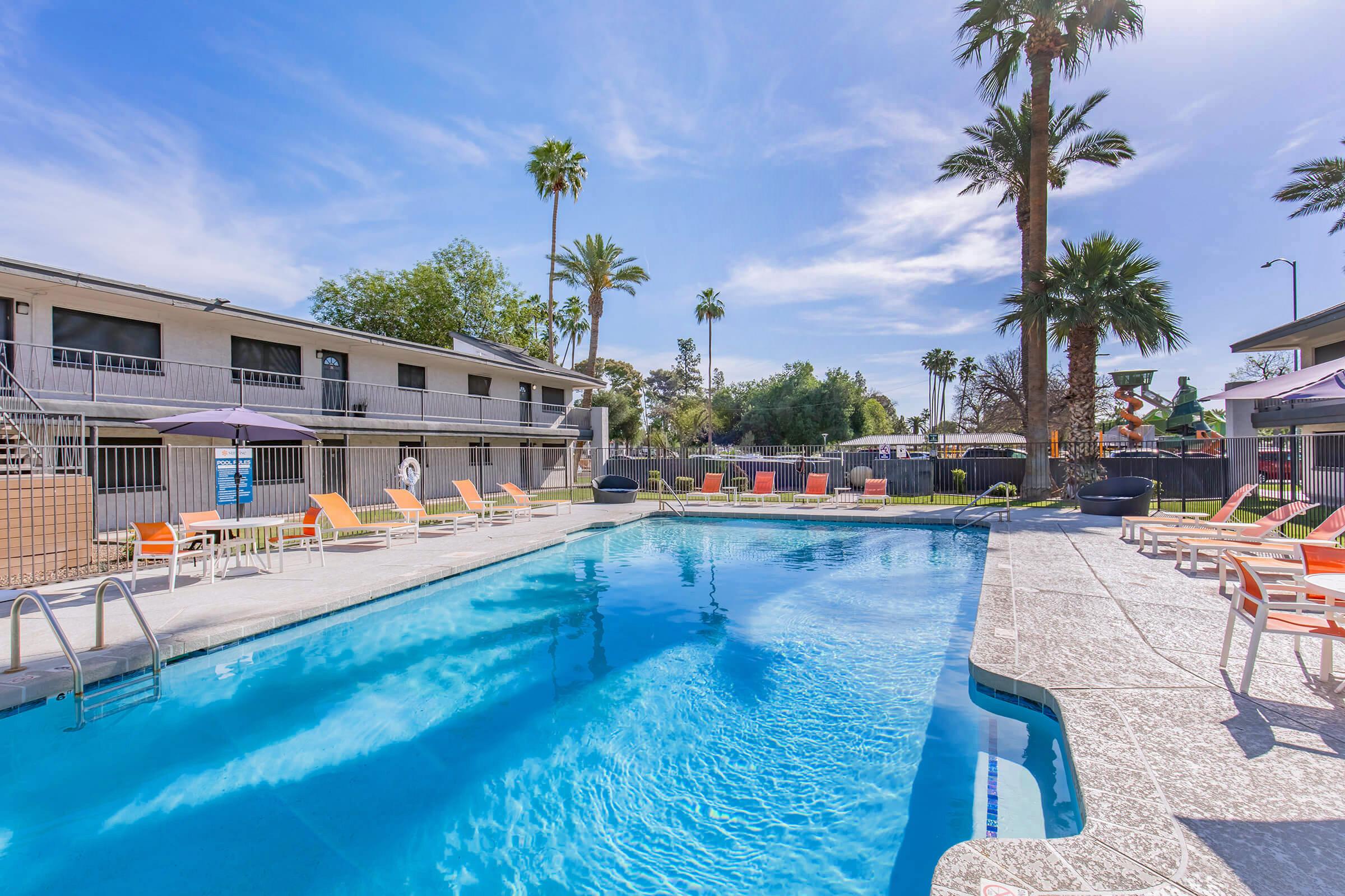 A clear blue swimming pool surrounded by lounge chairs under palm trees, with an apartment building in the background. The pool area is sunny, featuring a few umbrellas and a slide, creating a relaxing outdoor atmosphere.