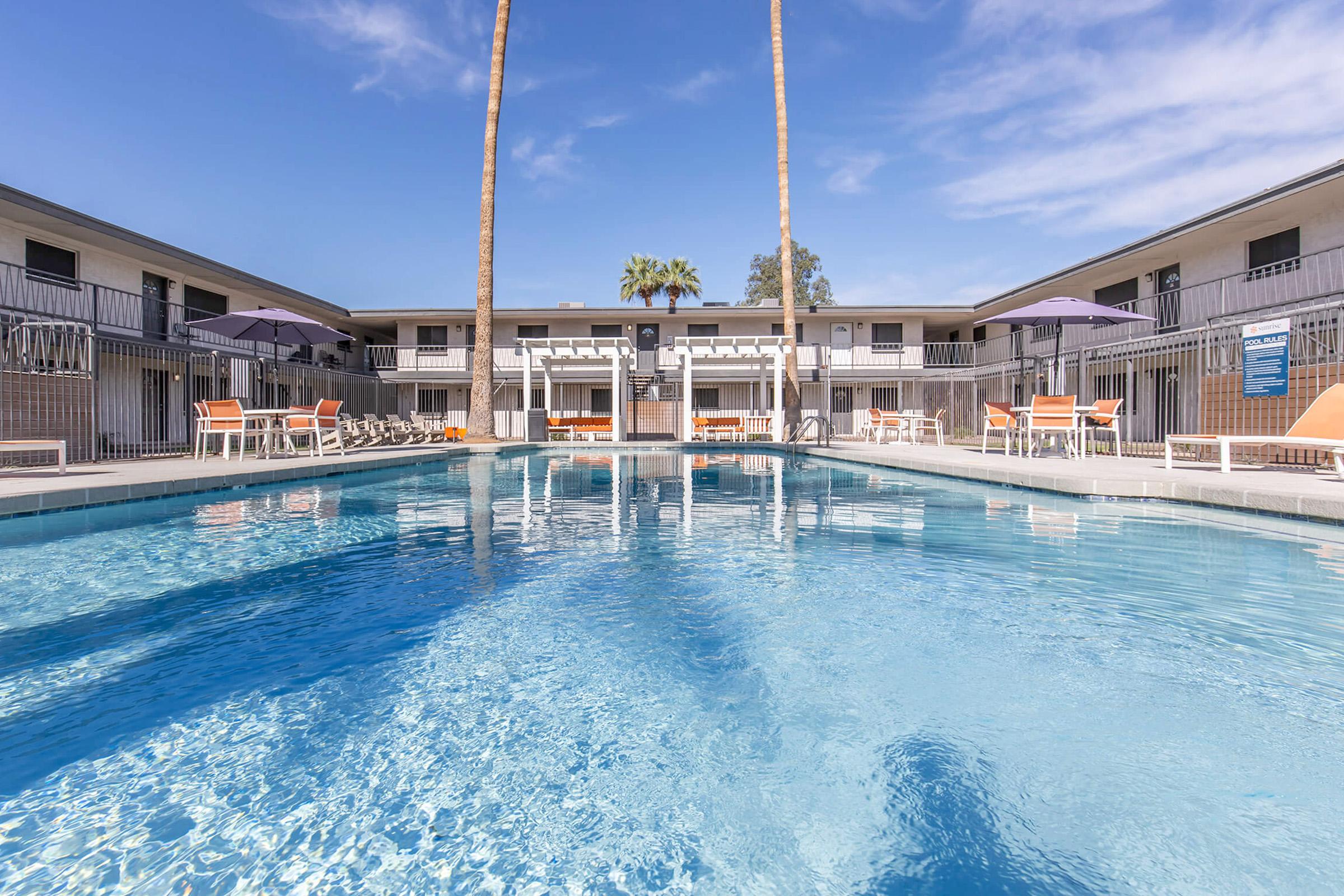 A clear blue swimming pool surrounded by lounge chairs and umbrellas, with palm trees in the background. The pool area is flanked by two-story buildings, creating a relaxing atmosphere under a bright blue sky.