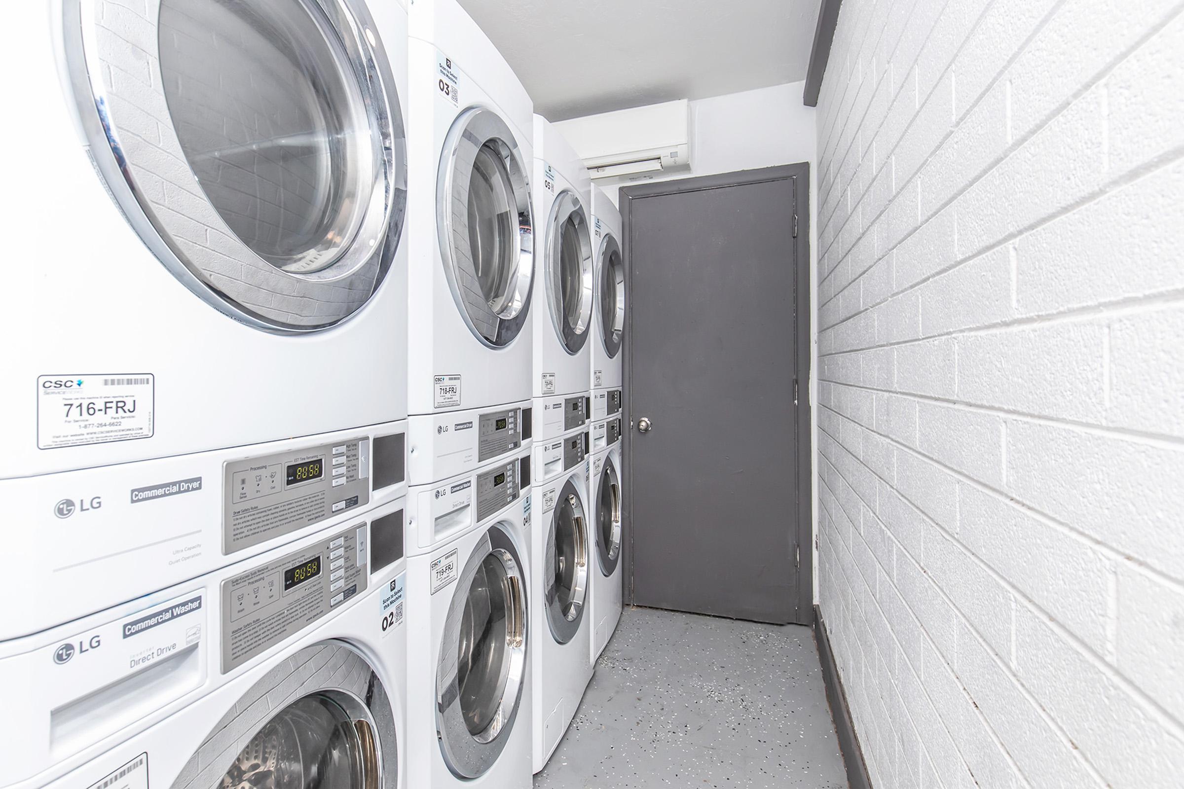A clean laundry room featuring stacked LG washer and dryer units against a white brick wall. There is a gray door on the right side, and the floor is light gray. The area appears well-lit and organized, suitable for laundry activities.