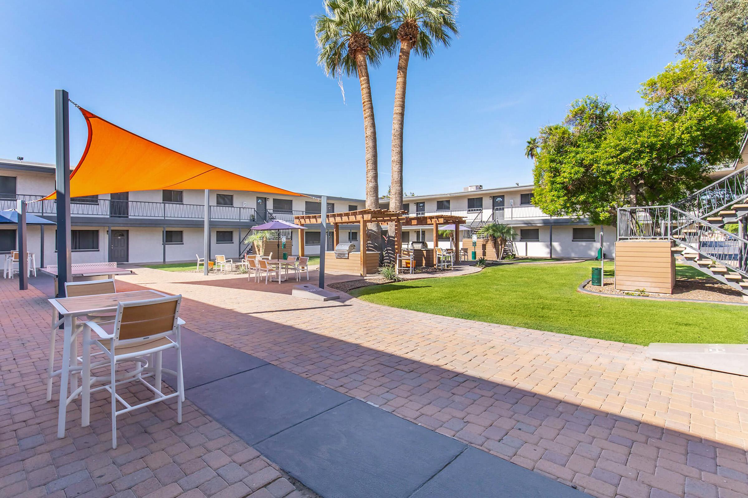 A sunny courtyard area featuring palm trees, orange shade sails, and picnic tables. Surrounding buildings are visible, and there are green lawns and seating areas, creating a relaxed outdoor space for leisure or gatherings.