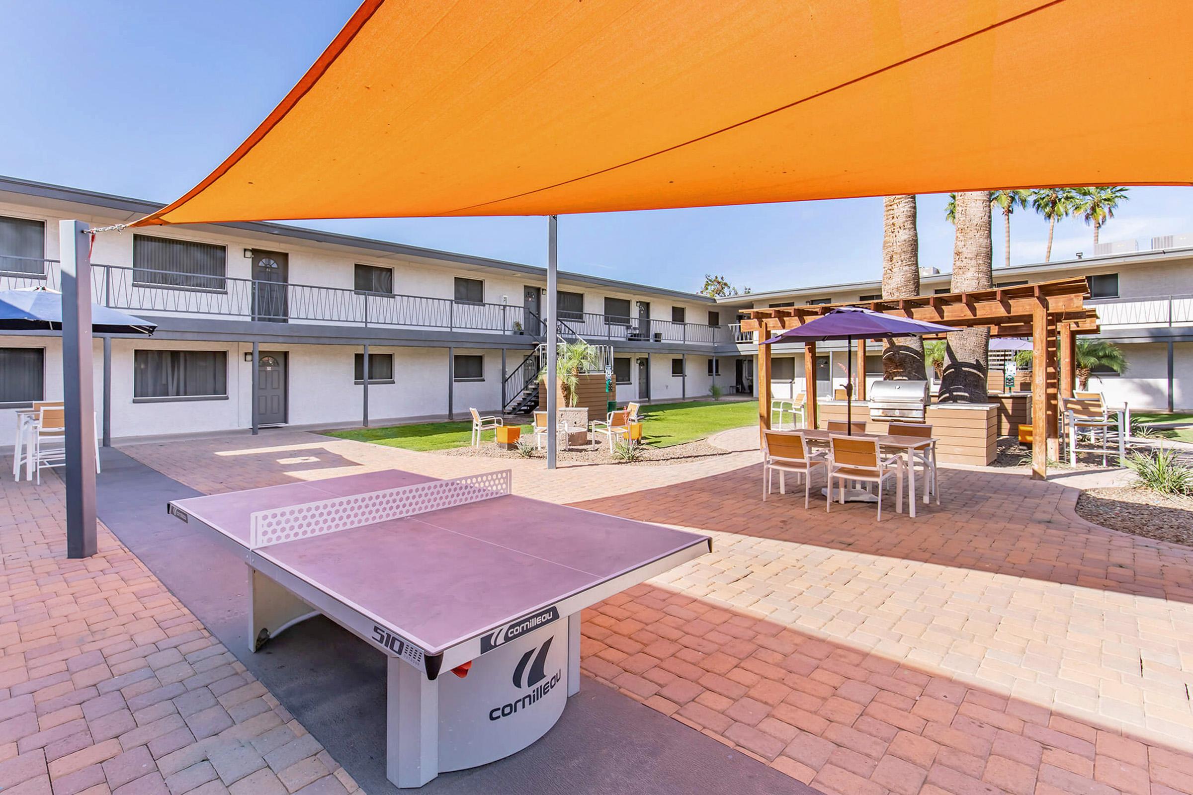 A courtyard with a ping pong table under a large orange shade sail. Surrounding the table are patio furniture, green grass, and palm trees, set against a backdrop of two-story white buildings. The area is designed for relaxation and socializing, with a pleasant outdoor atmosphere.