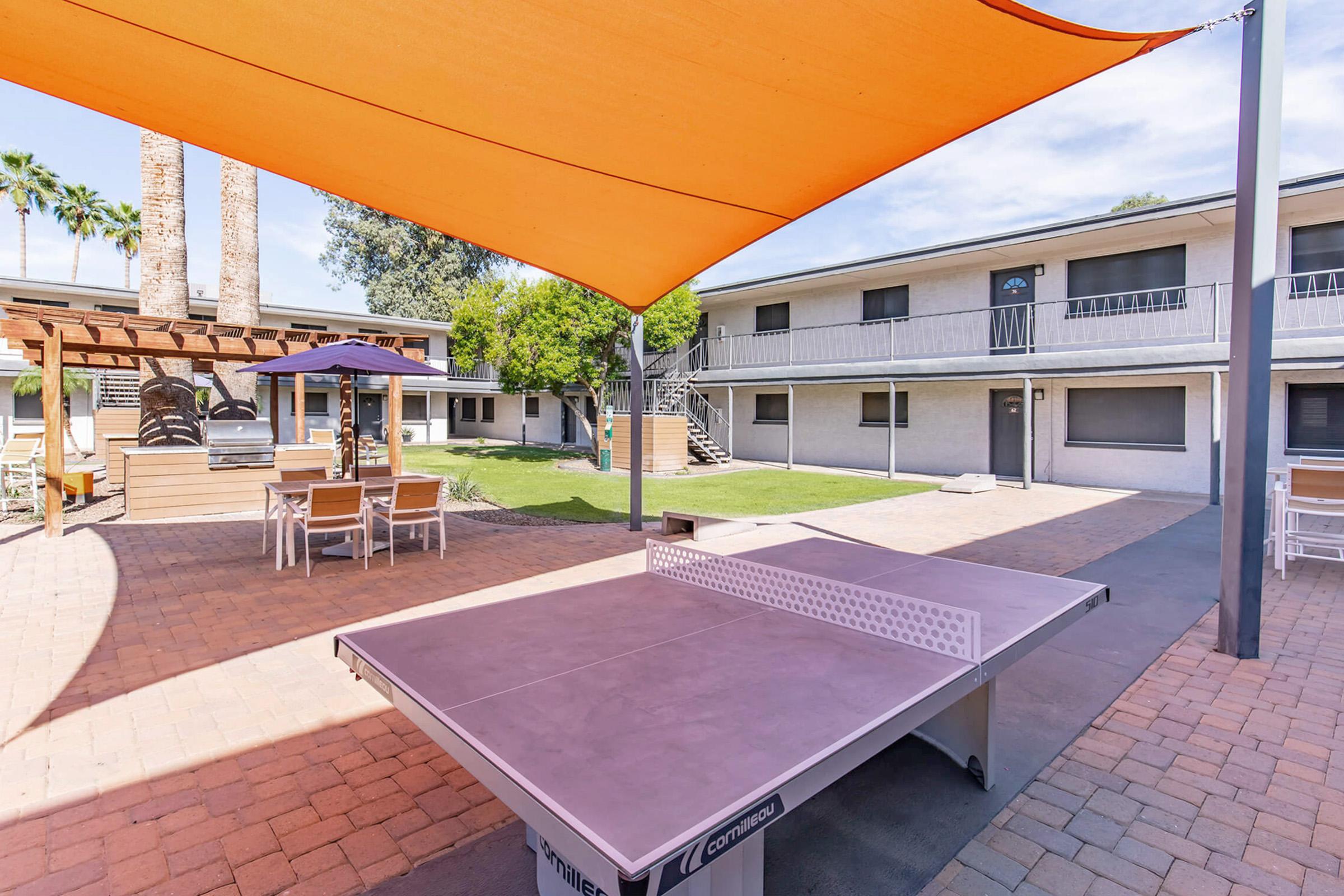 A sunlit courtyard featuring a ping pong table under a large orange shade sail. Surrounding the area are several outdoor seating arrangements, including tables and chairs, with palm trees and two-story buildings in the background. The setting offers a relaxed environment for socializing and recreation.