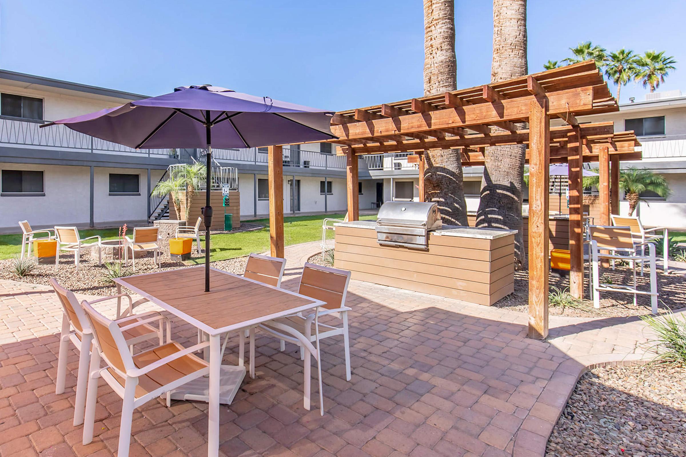 A cozy outdoor patio area featuring a wooden pergola with a purple umbrella, a table with chairs, and a grill surrounded by palm trees and well-maintained landscaping. The background includes two-story apartment buildings, creating a welcoming space for relaxation and gatherings.