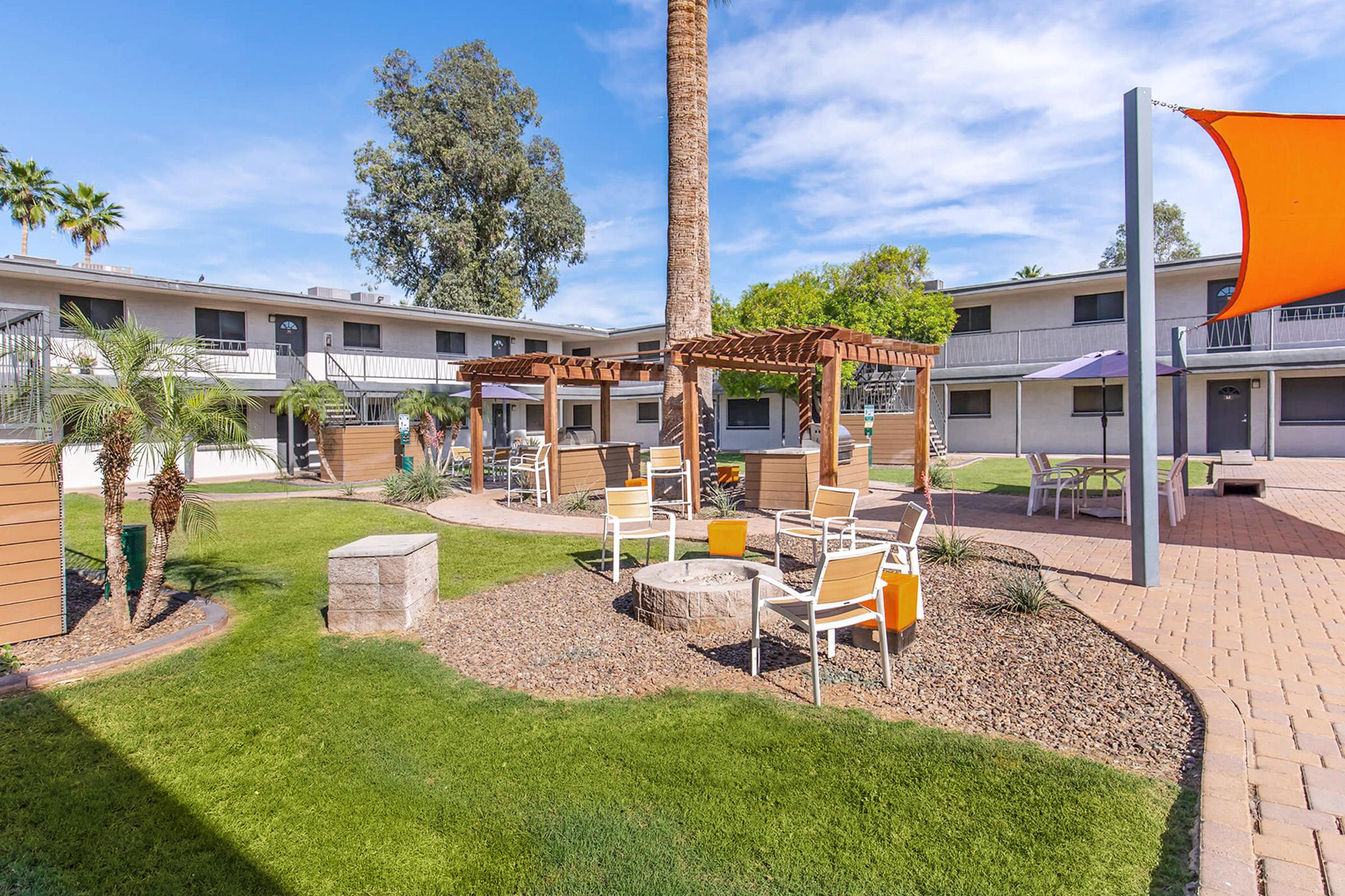 A landscaped outdoor area featuring a circular seating arrangement around a fire pit, surrounded by palm trees and decorative plants. Bright orange accents are present in the chairs. In the background, a two-story building and shaded structures provide a relaxing atmosphere. Skies are clear with scattered clouds.