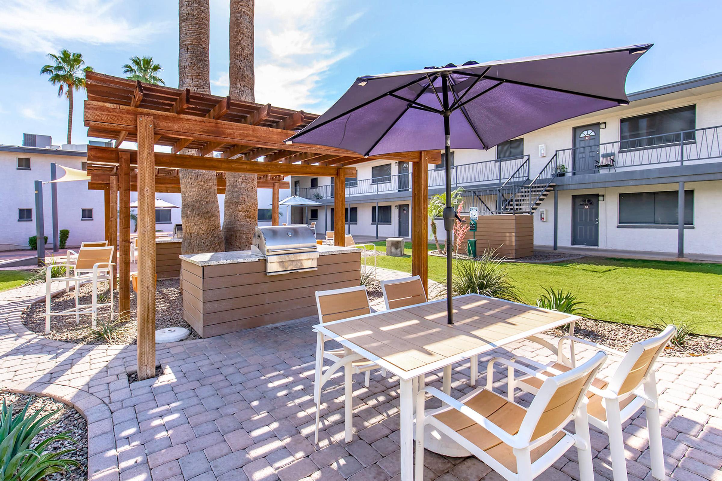 Outdoor seating area with a picnic table and umbrella, surrounded by palm trees and green grass. A barbecue grill is positioned nearby under a wooden pergola. In the background, there are white apartment buildings with stairs leading to the upper levels. Clear blue sky above creates a bright atmosphere.