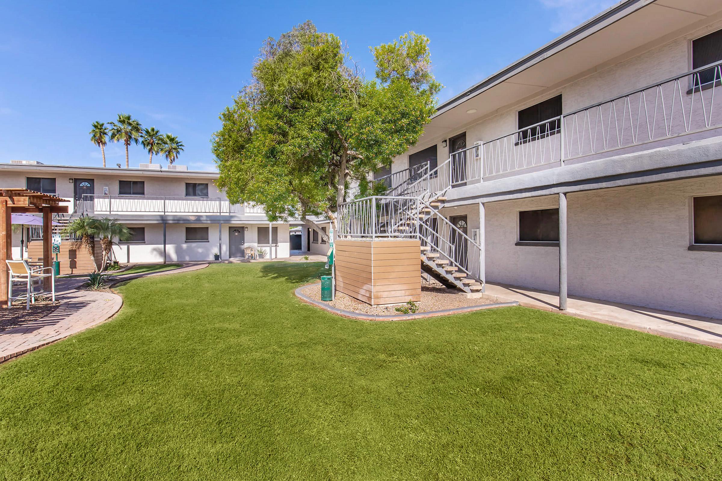 A landscaped courtyard featuring green grass, palm trees, and a spiral staircase leading to an upper level of an apartment complex. The building has a light-colored exterior with railings on the balconies, and there is a small play area nearby. The scene is bright and sunny, showcasing a residential environment.