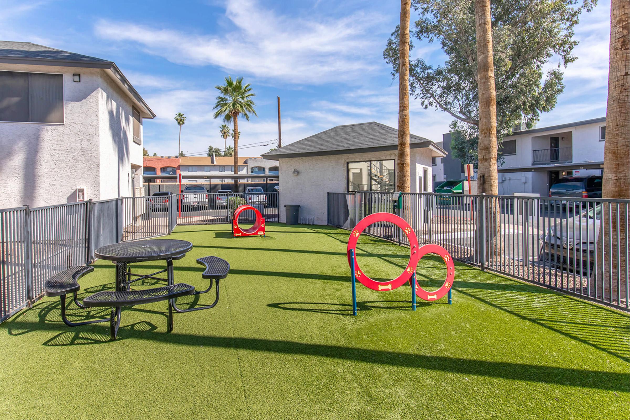A well-maintained outdoor area featuring a green turf lawn, a picnic table, and interactive red play equipment. The setting is surrounded by palm trees and includes nearby buildings, creating a welcoming space for relaxation and play. Ideal for pets or families enjoying the outdoors.