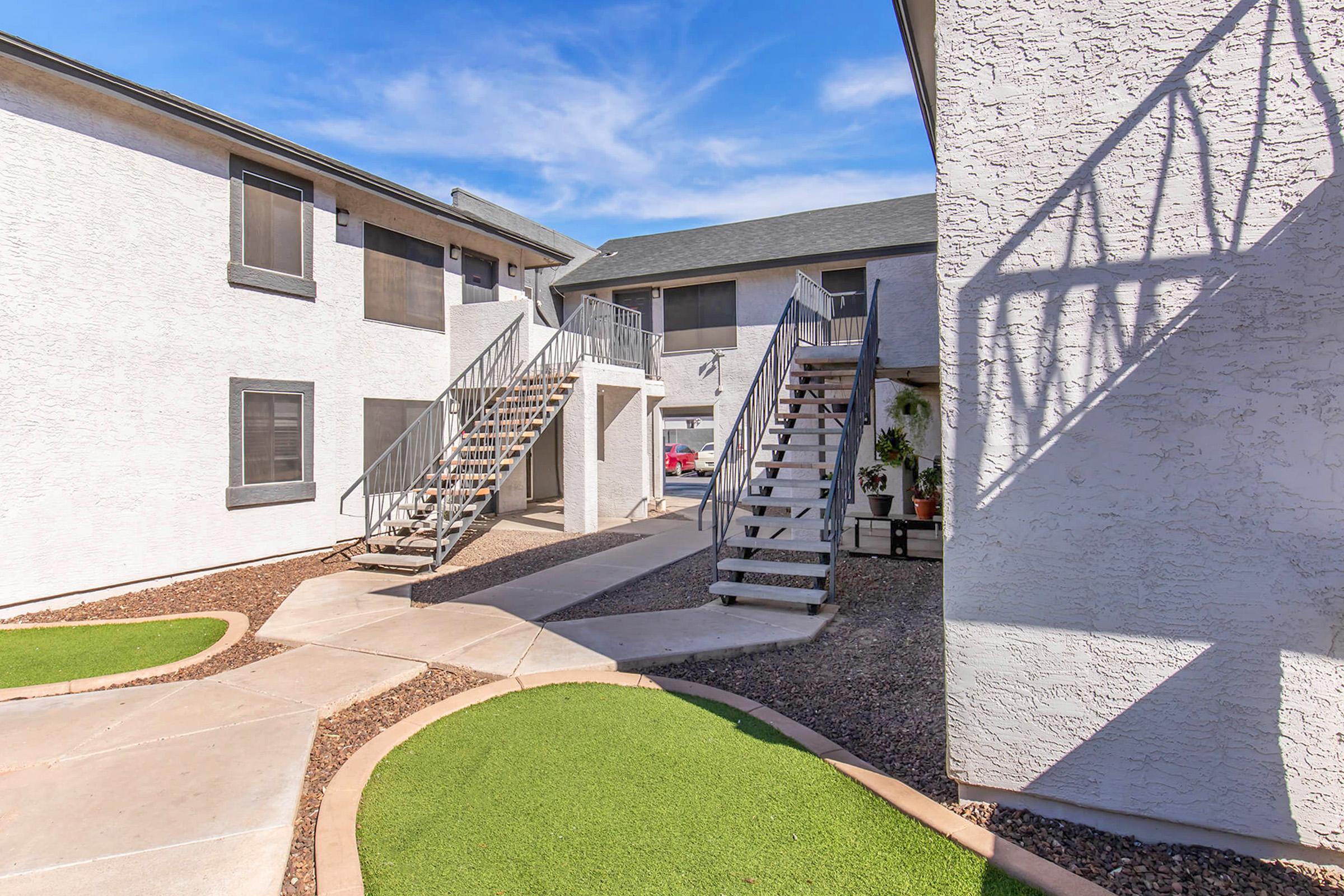 Two apartment buildings with white exteriors are visible, featuring outdoor staircases leading to second-floor units. A landscaped area with artificial grass and decorative stones is in the foreground, while a clear blue sky is seen above.