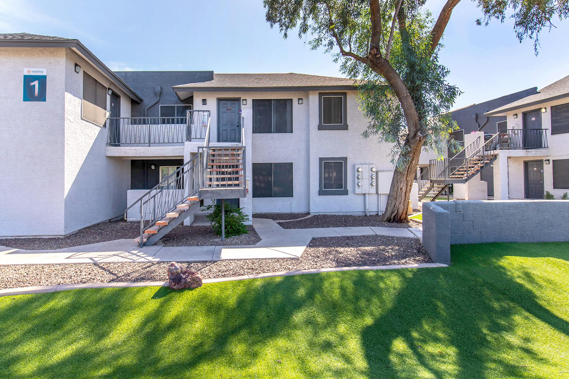 Multi-story apartment building with a stairway leading to the entrance. The exterior features light-colored walls and dark trim. Surrounding the building is a landscaped area with green grass and trees, providing a well-maintained atmosphere. Clear blue sky visible above the structure.