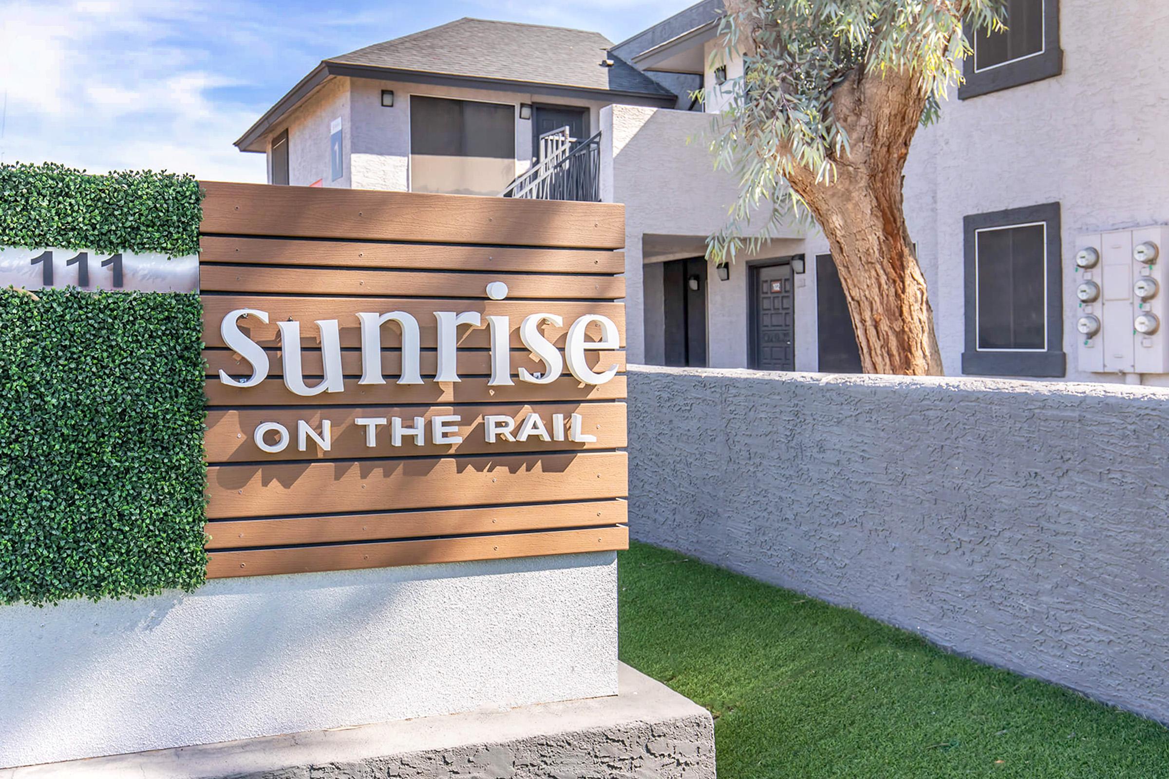 Sign for "Sunrise on the Rail" displayed on a wooden panel with greenery. The sign is positioned in front of a multi-unit residential building featuring a tree and exterior utility meters nearby. The setting is bright and inviting, suggesting a modern living environment.