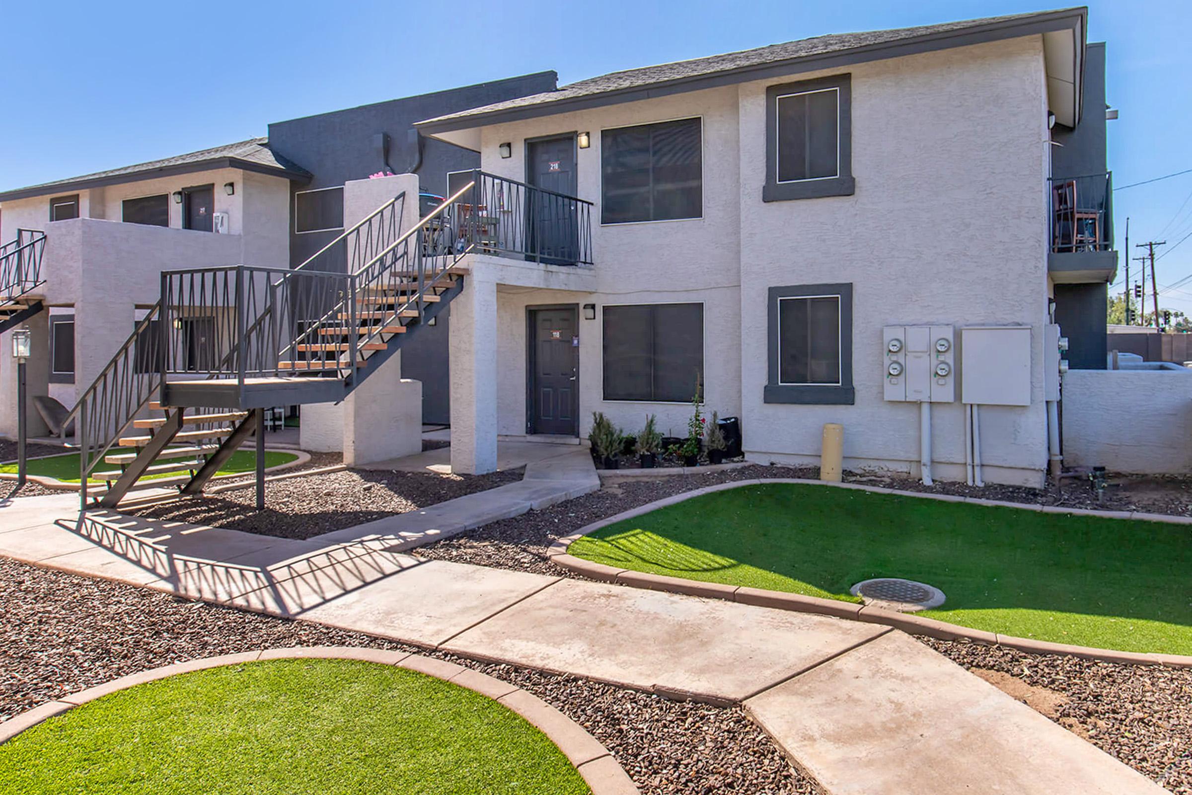 Apartment building featuring a two-story structure with a gray and white facade. A staircase leads to the second floor, while landscaped areas with artificial grass and pebbles surround the building. Utility meters are visible on the side, and the sky is clear with bright sunlight.
