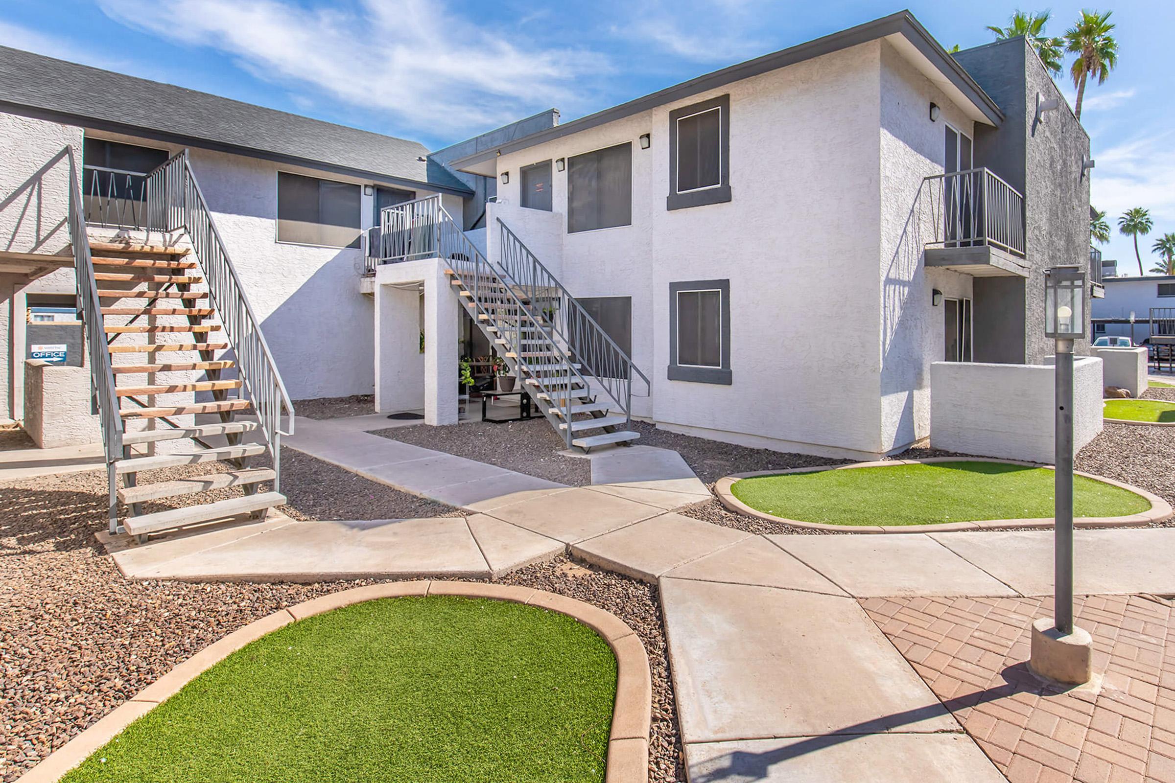 A view of a multi-unit apartment complex featuring two buildings with staircases, pathways, and landscaped areas. The exterior is painted white, with a gray roof. Decorative stones and small patches of grass are arranged around the walkways, creating a welcoming outdoor space.
