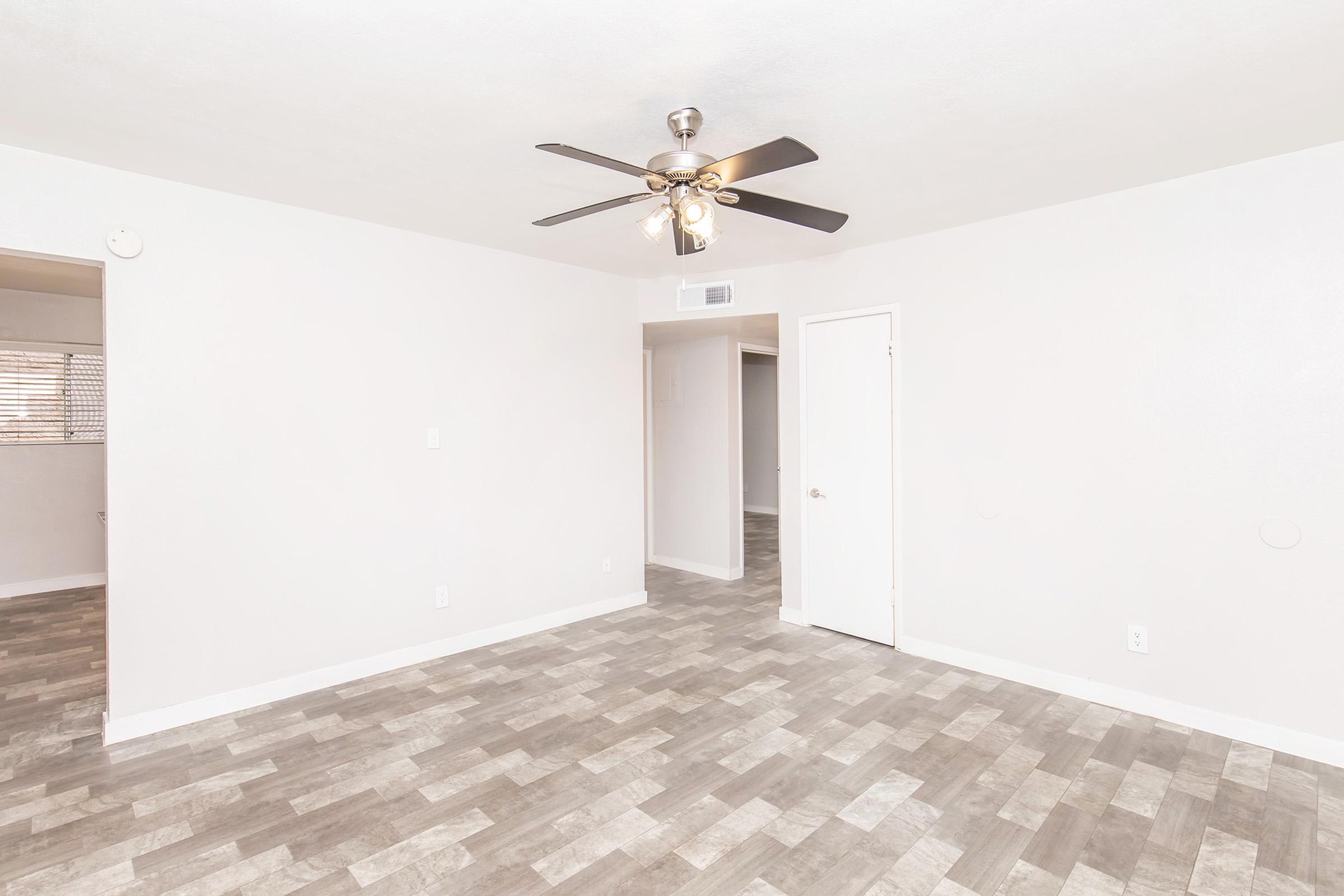 Empty interior space of a room featuring a light-colored wall, a ceiling fan with multiple blades, and a gray tiled floor. An open door leads to another room, and there is a window obscured by blinds in the background. The overall atmosphere is bright and uncluttered.