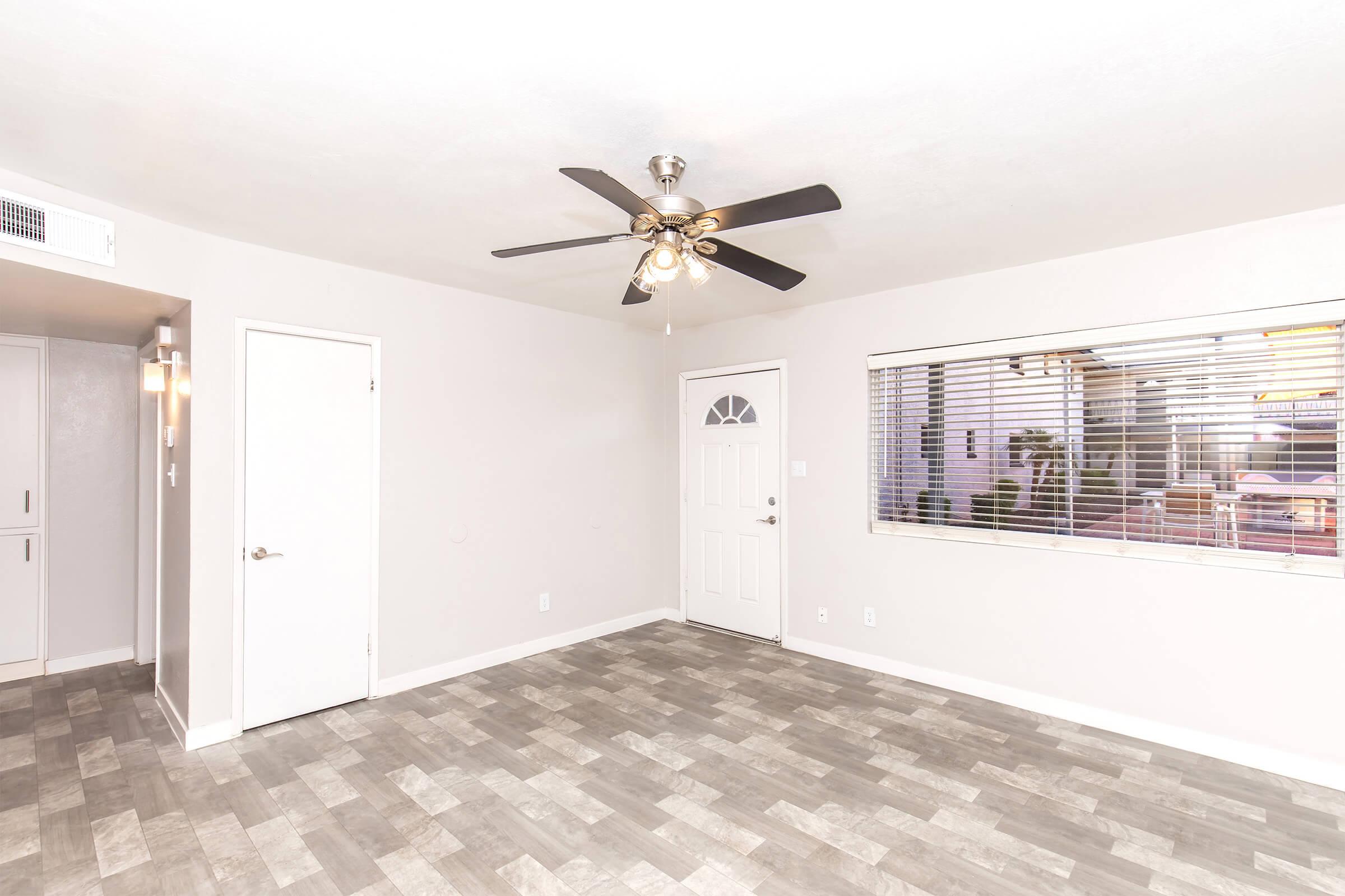 An empty room featuring a ceiling fan, a door, and a window with blinds. The flooring is a light-colored tile pattern. The walls are painted in a neutral color, creating a clean and spacious atmosphere.