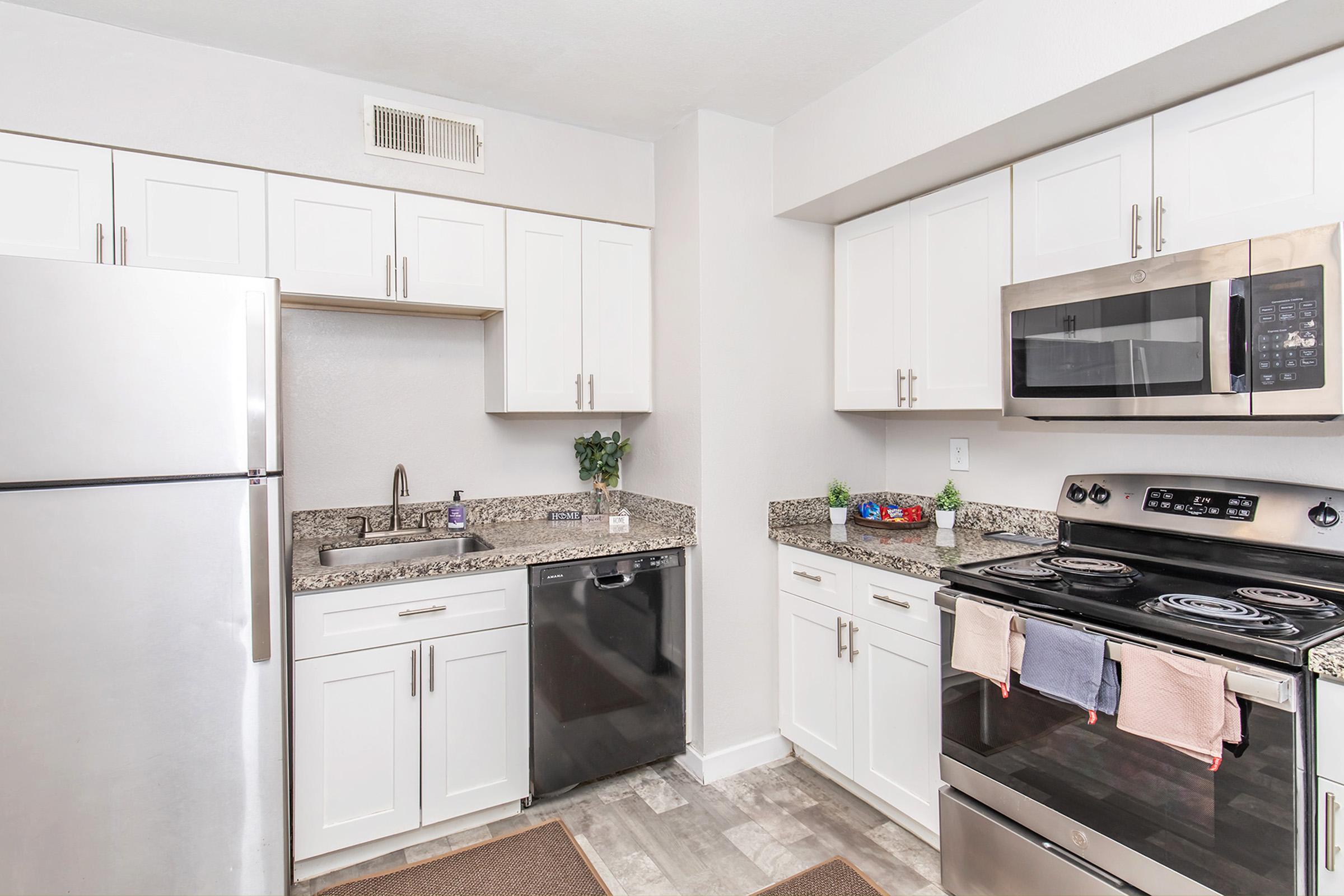 Modern kitchen with white cabinets, granite countertops, and stainless steel appliances. Features a refrigerator, microwave, stove, and dishwasher. The space is bright with neutral walls and a small decorative plant on the countertop. Cookware and a dish towel are visible on the stove.