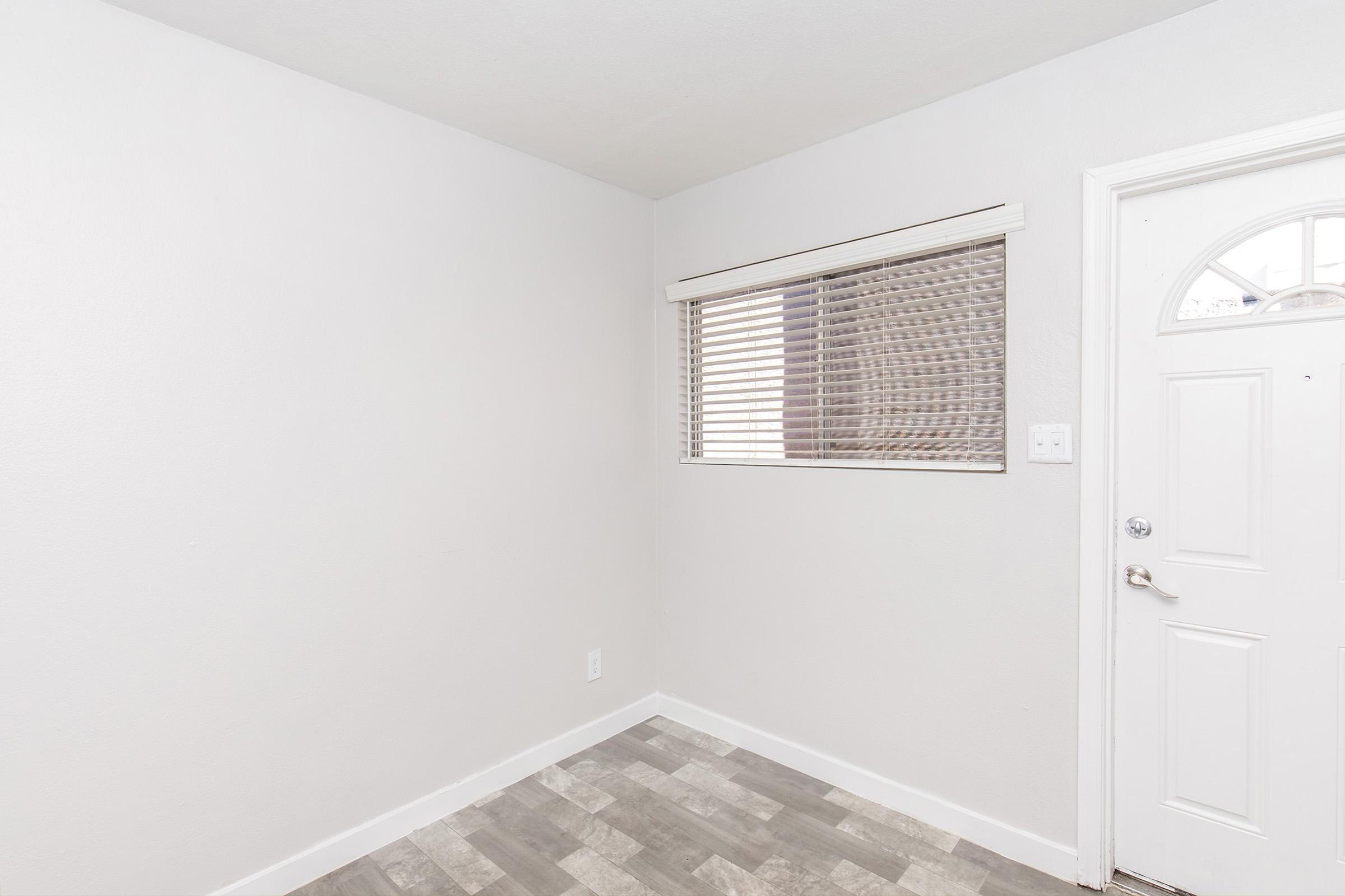 A minimalist room with light gray walls and a window covered by horizontal blinds. The floor features a patterned tile design in shades of gray. A white door on the right leads to the outside, creating a bright and airy atmosphere. The space is uncluttered and ready for decoration.