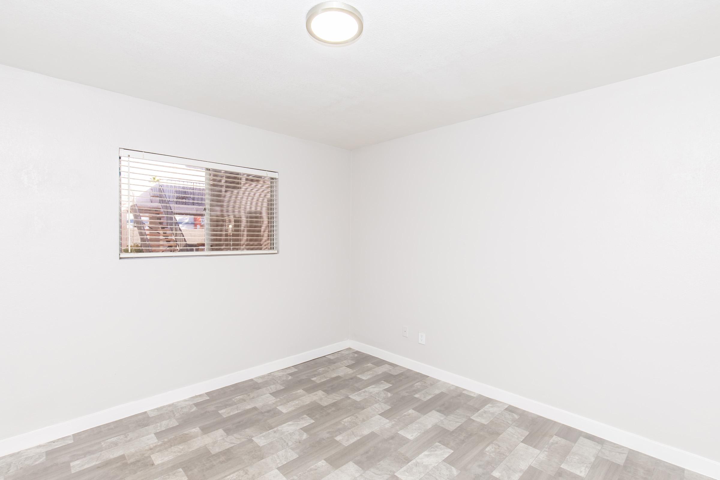 A lightly colored empty room with gray tile flooring. There is a window with horizontal blinds on one wall, showing a glimpse of the outdoors. The walls are painted in a neutral shade, creating a spacious and bright atmosphere.