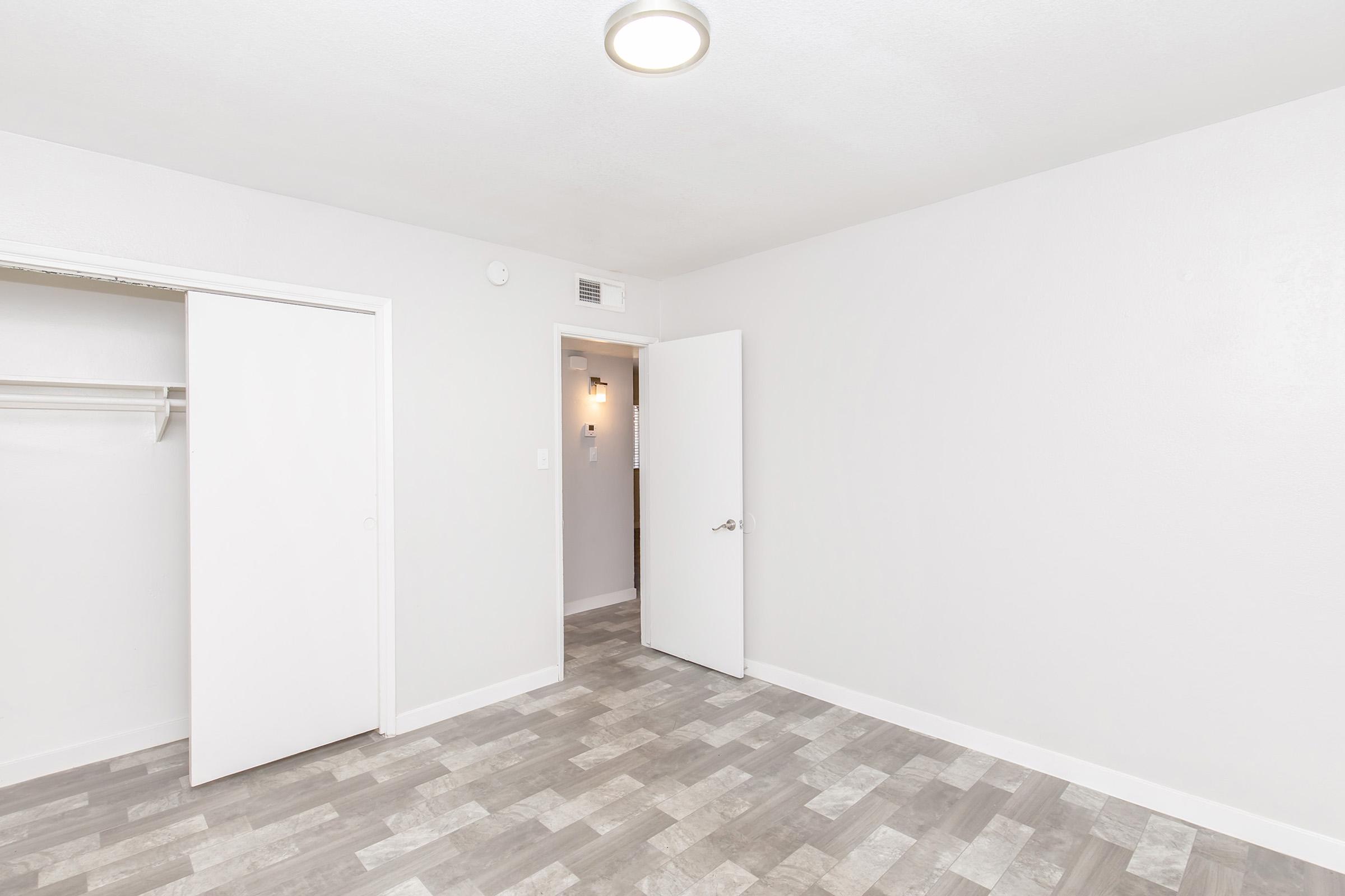 A vacant room with light gray walls, a circular ceiling light, and a floor featuring wood-like tiles. A closet with sliding doors is on the left, and an open door leads to another area. The room has a clean, modern appearance and is well-lit.