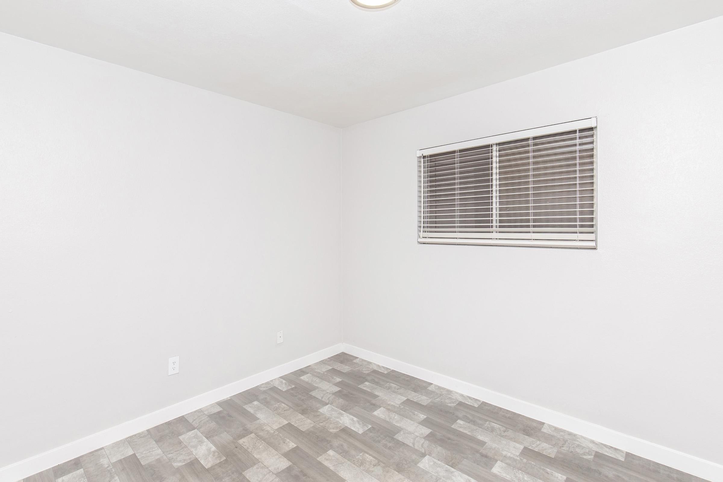 Empty room with light gray walls and a window featuring white blinds. The flooring consists of light gray wood-like tiles. The space appears clean and minimalistic, suitable for various uses.