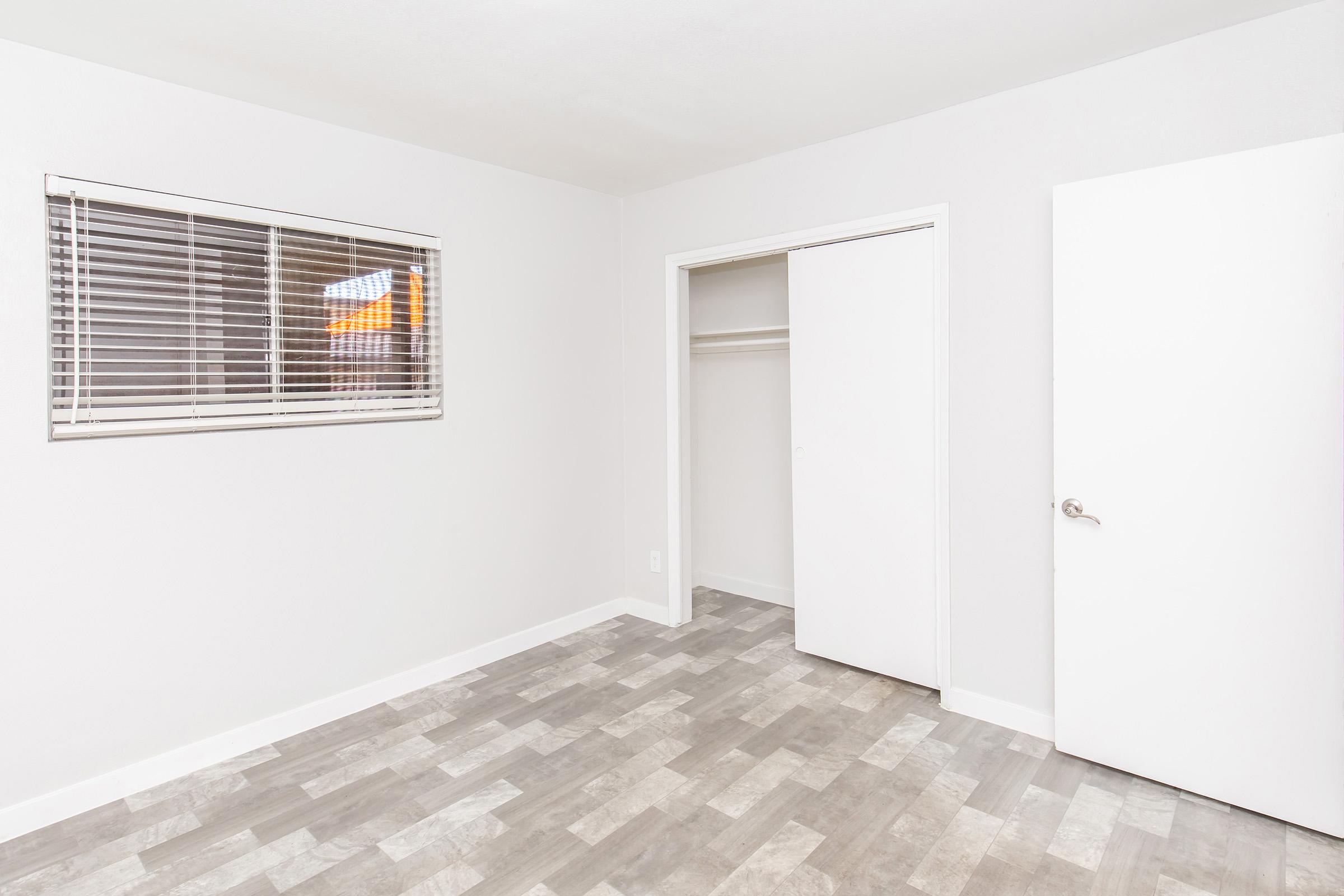 A simple, empty room with light gray walls and a window featuring horizontal blinds. The floor is covered in light gray and beige tiles. There is a closet with sliding doors on one side and a plain white door on the other. The room has a bright and spacious feel.