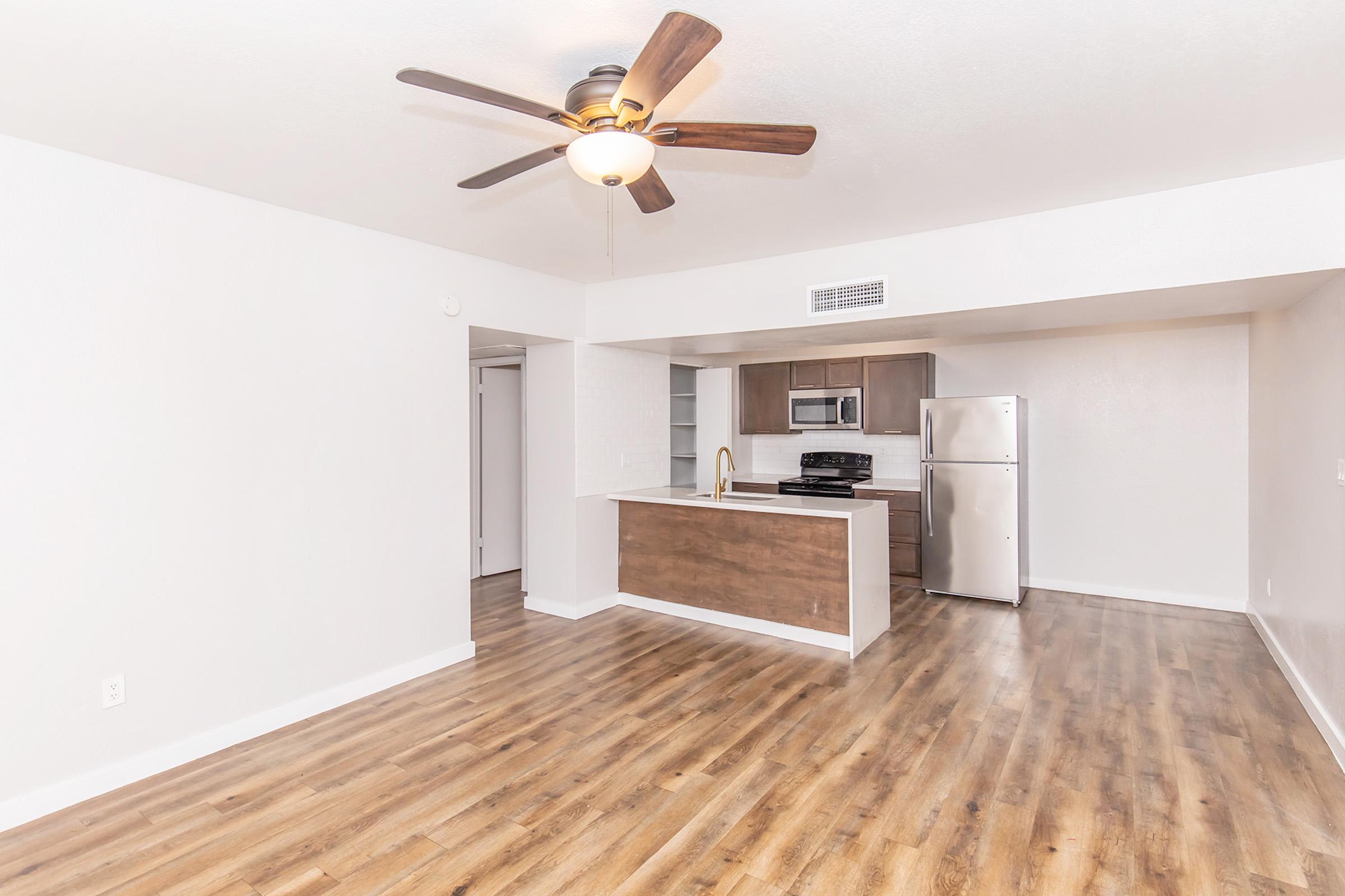 Spacious living area featuring a modern kitchen with stainless steel appliances, a ceiling fan, and hardwood-style flooring. The open layout connects the kitchen and living space, with neutral walls and ample natural light. Complete with a doorway to a bedroom and a sleek refrigerator.