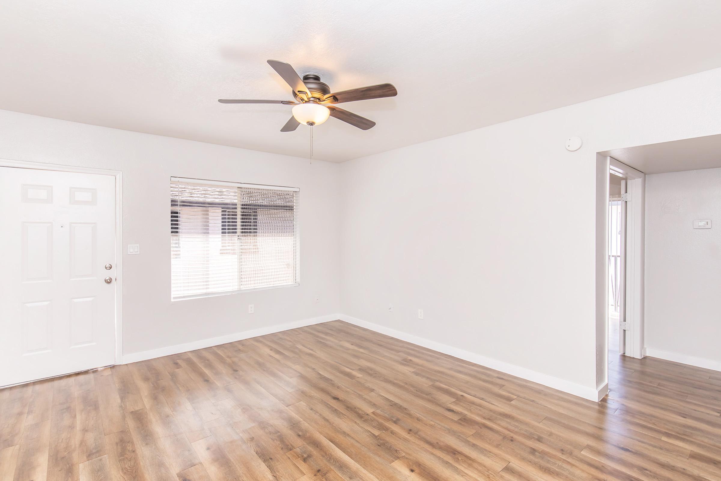 A bright, empty living room featuring light gray walls, a ceiling fan with a light fixture, and a large window with blinds. The flooring is a warm wood laminate, and a front door is visible on the left, with an open hallway to the right leading to another room. The space is well-lit and inviting.